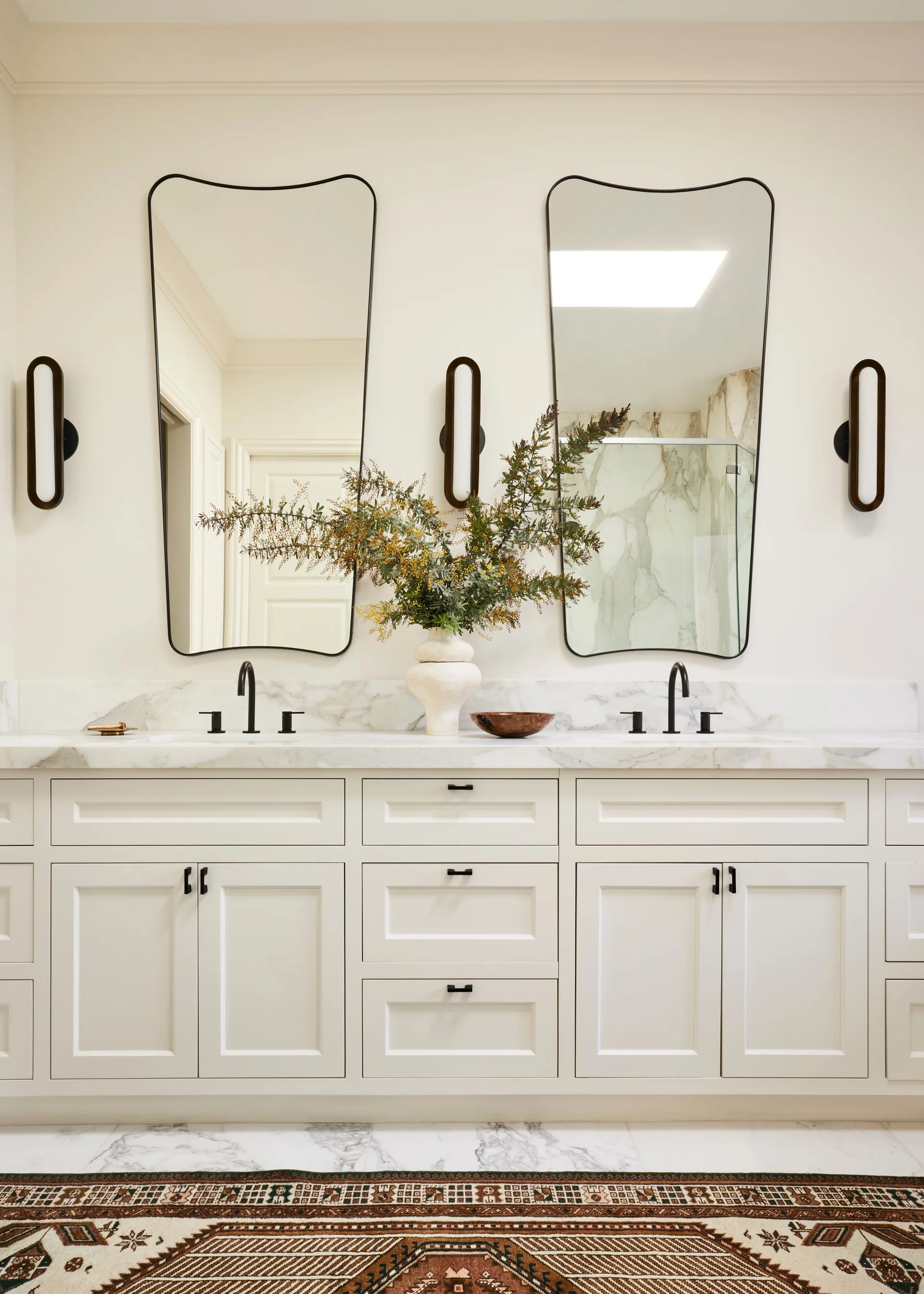 A white double vanity bathroom features two asymmetrical black-rimmed mirrors, pill-shaped sconces, marble countertops, and a patterned rug.