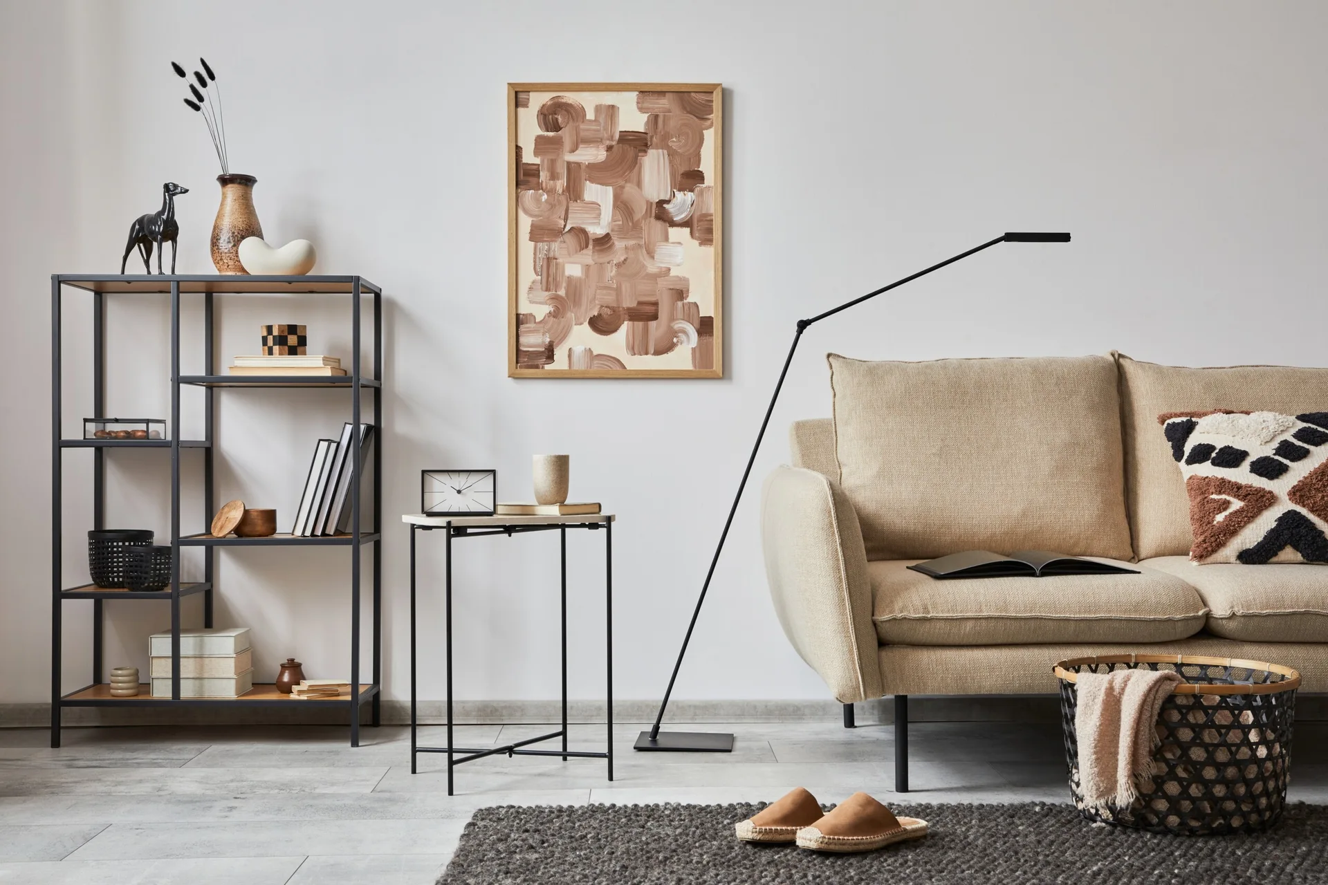 A minimalist living room featuring a beige fabric sofa, a black metal shelving unit, a slim side table, and abstract wall art.