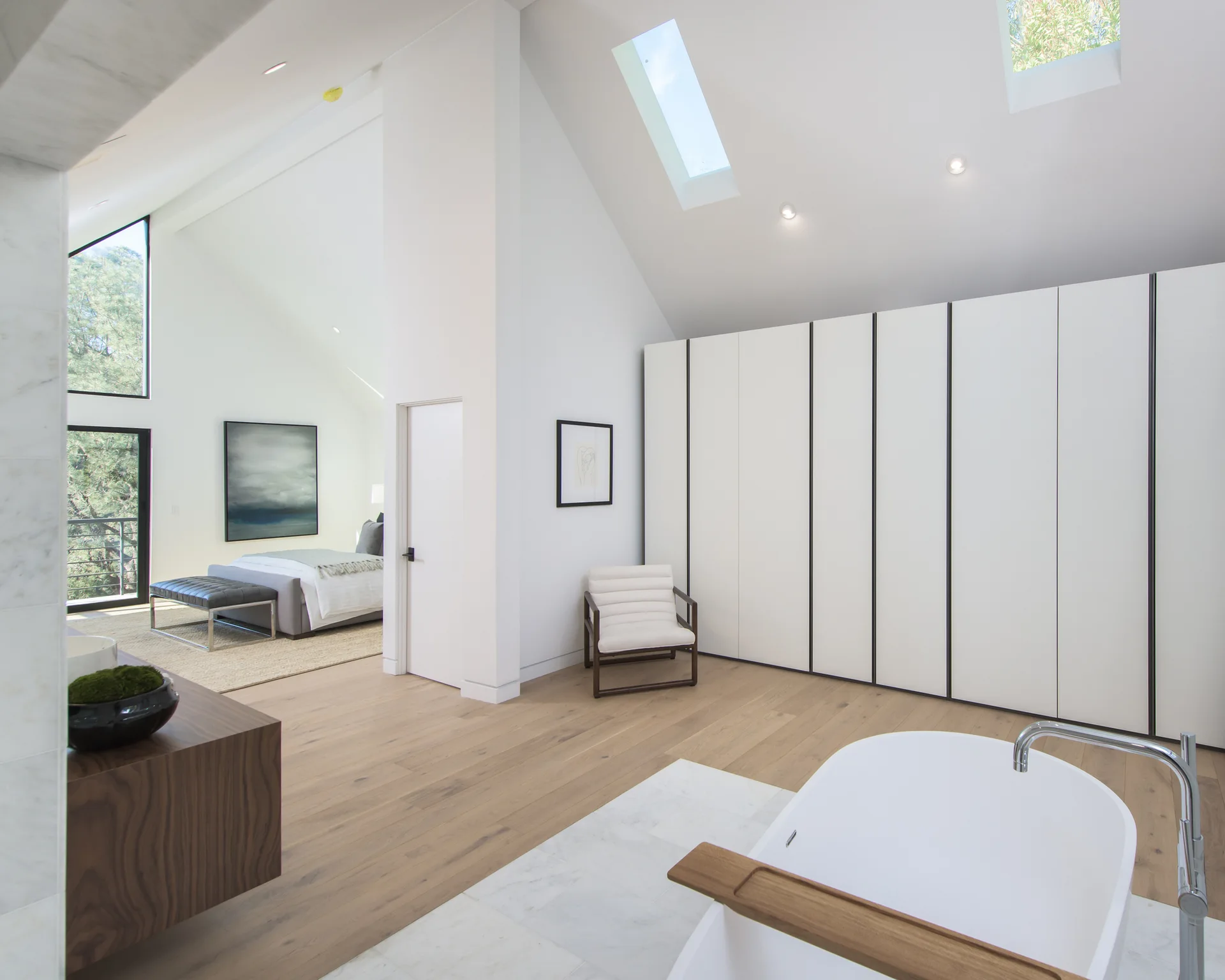 A modern primary suite featuring a white soaking tub, light wood floors, vaulted ceilings with skylights, and a large white wardrobe cabinet.