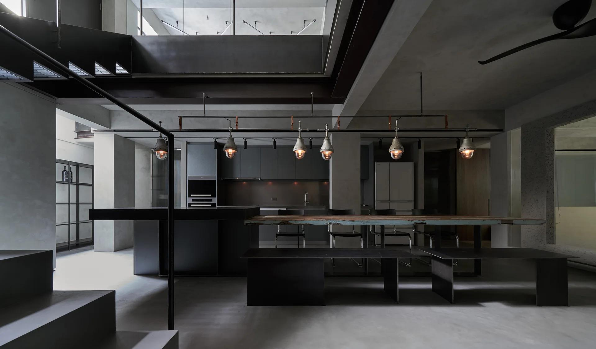 An industrial kitchen features gray concrete walls, a raw wood dining table connected to a dark island, and a row of low-hanging pendant lights.