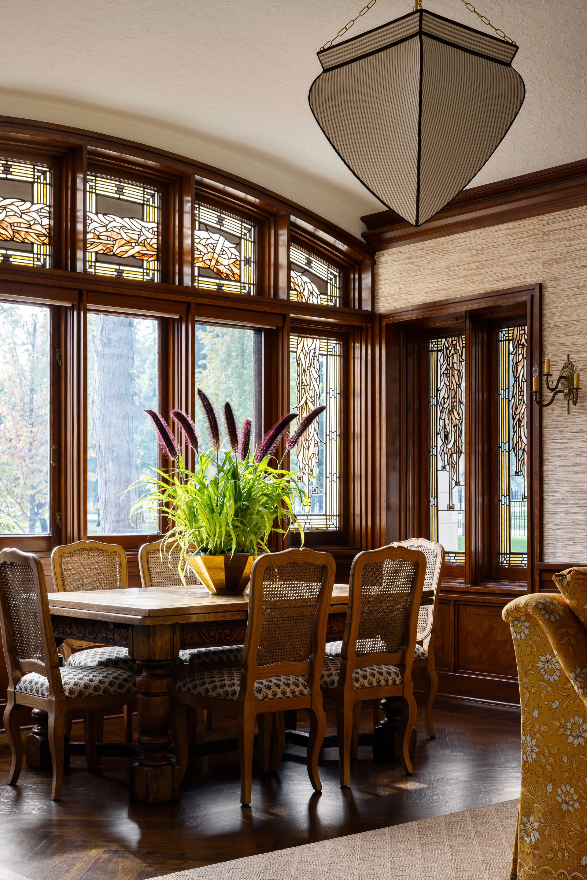 A traditional dining room features a wooden table surrounded by cane-back chairs, set against dark wood trim and stained glass windows.