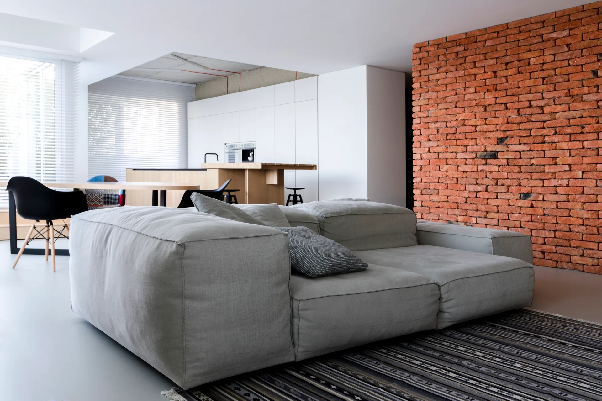 A modern open-plan living room with a large grey modular sofa on a striped rug next to a prominent orange brick feature wall.