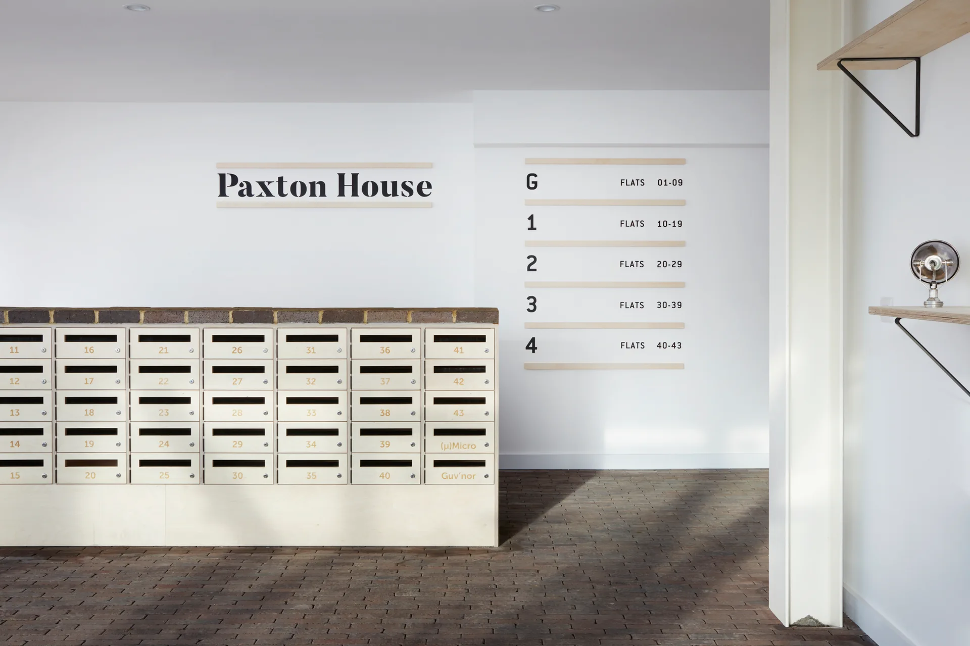 A minimalist apartment lobby features a custom light wood mailbox unit and matching wall directory set against white walls and a dark brick floor.