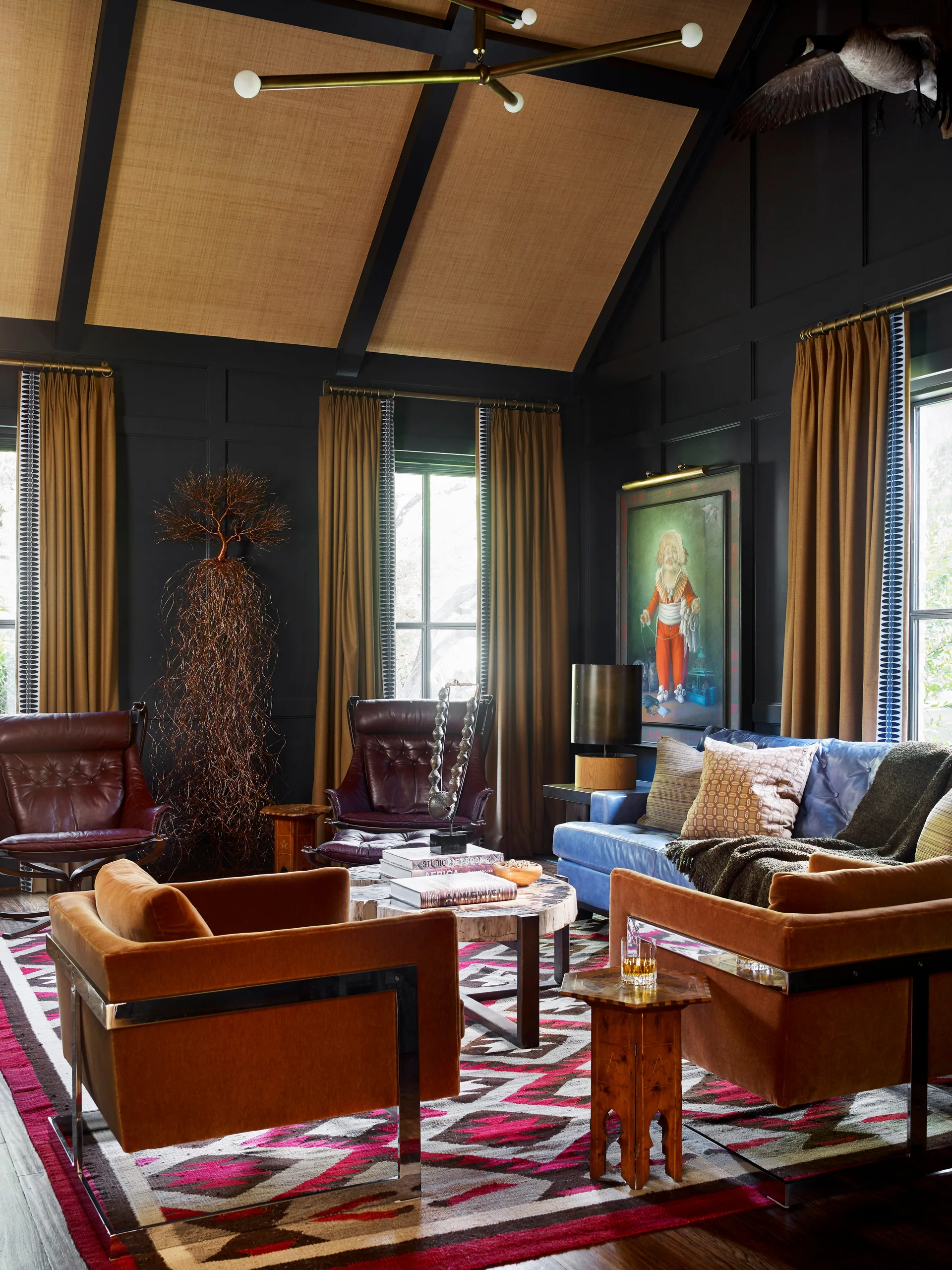 A moody living room features dark charcoal paneled walls, a vaulted ceiling, a blue leather sofa, rust-colored velvet armchairs, and a geometric rug.