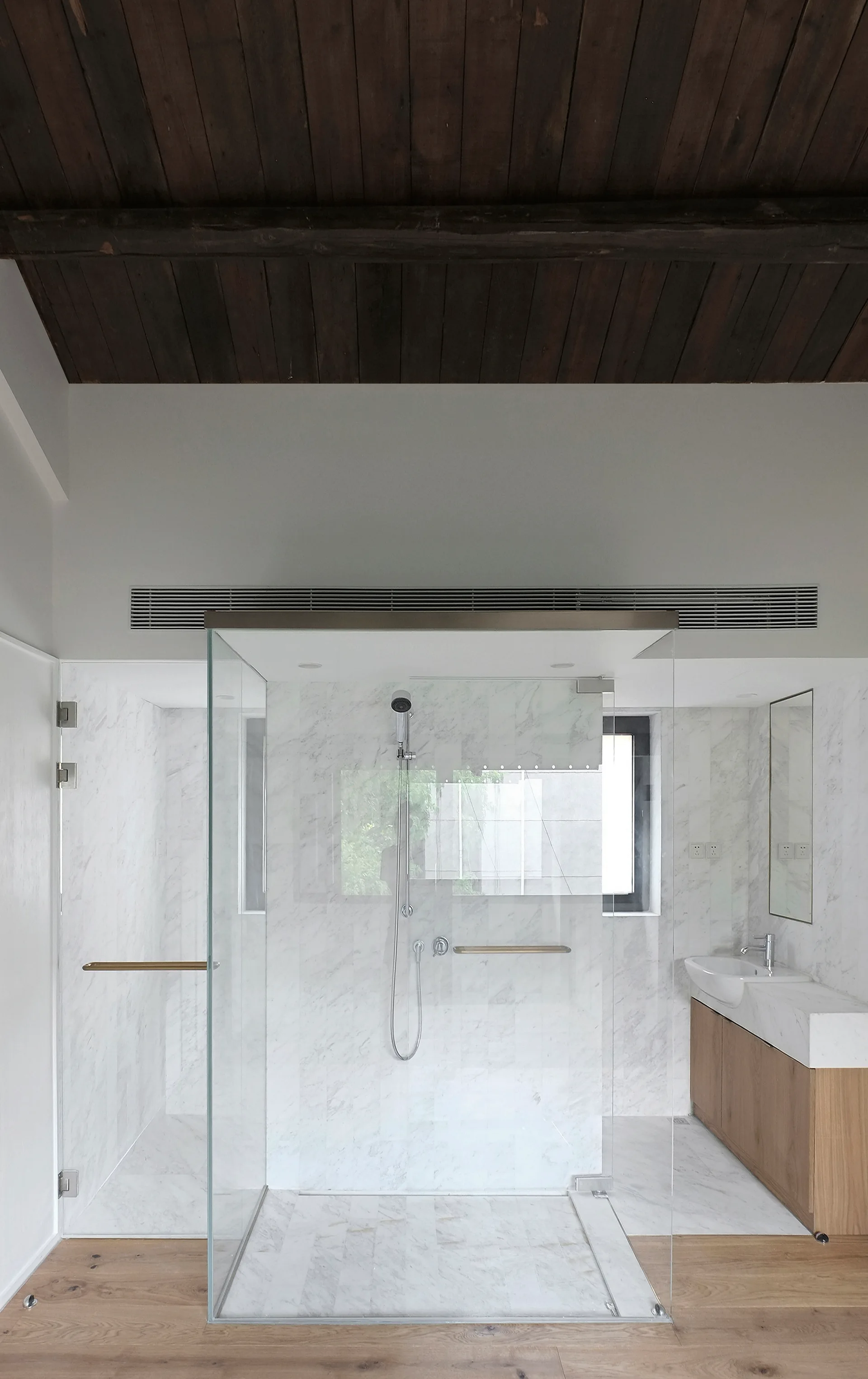 A minimalist bathroom featuring a glass walk-in shower with marble tile walls under a dark wood plank ceiling.