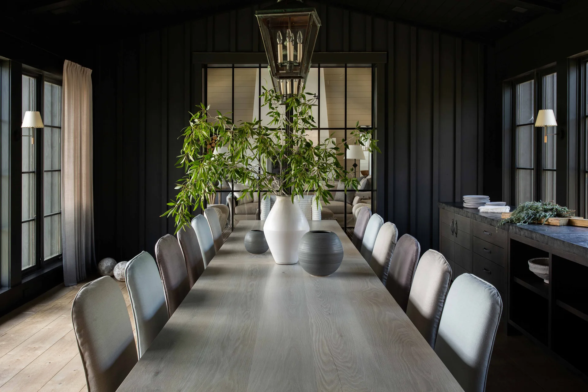 A long light wood dining table with grey chairs sits in a room with black vertical wood paneled walls and a metal glass partition.