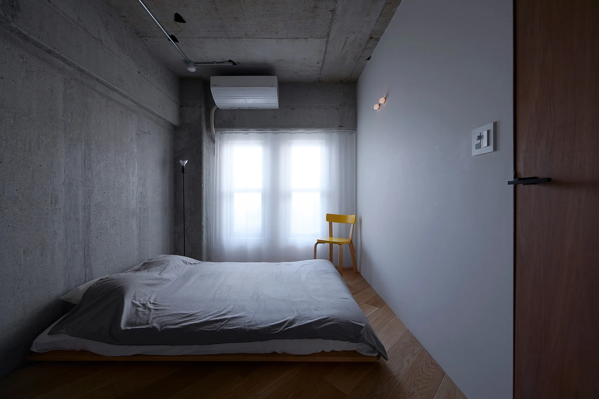 A bedroom featuring a low wooden platform bed on diagonal floors, set against raw grey concrete walls and a bright window with sheer curtains.