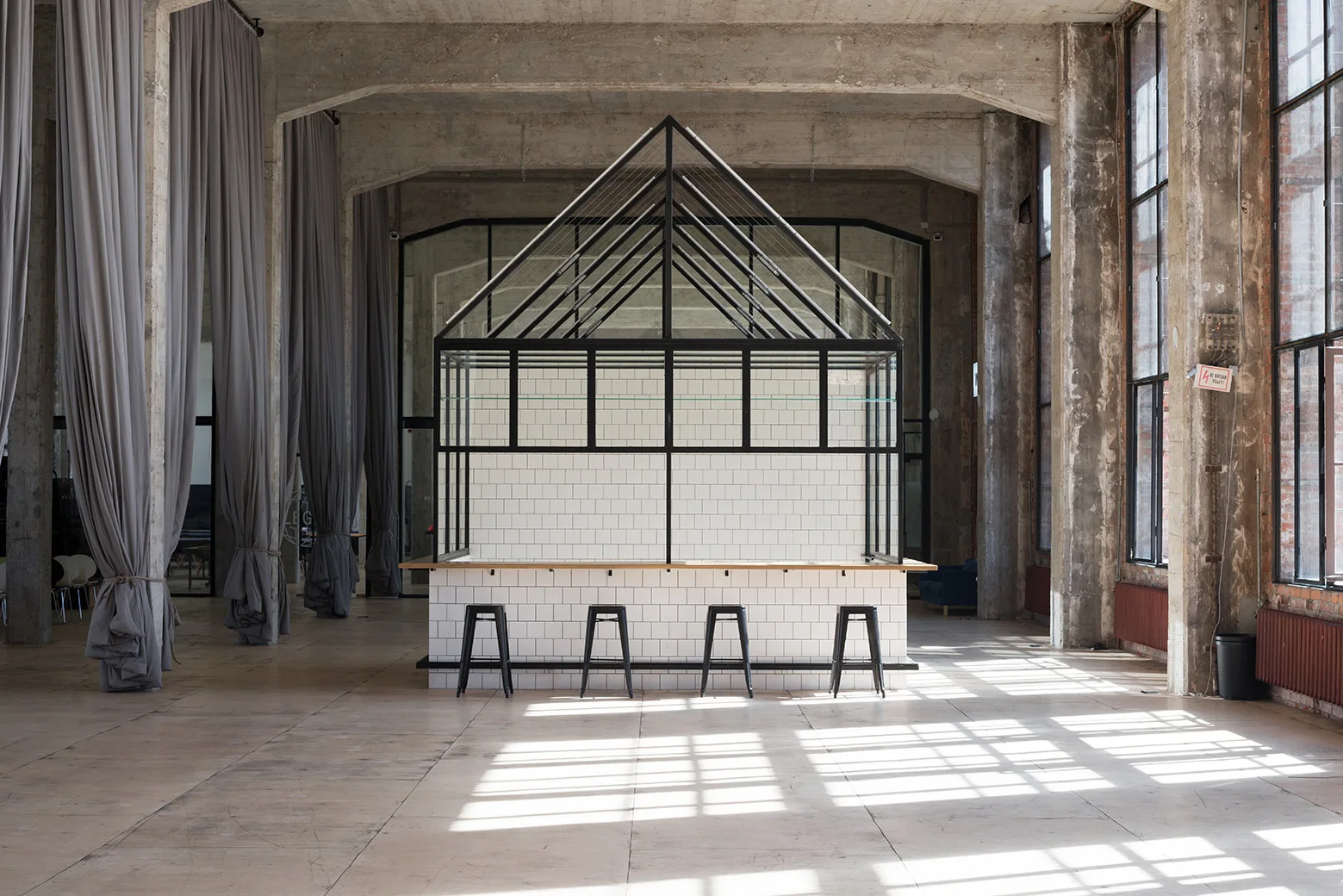 A central tiled bar with a gabled metal house frame stands on a concrete floor inside a spacious industrial room with tall windows.