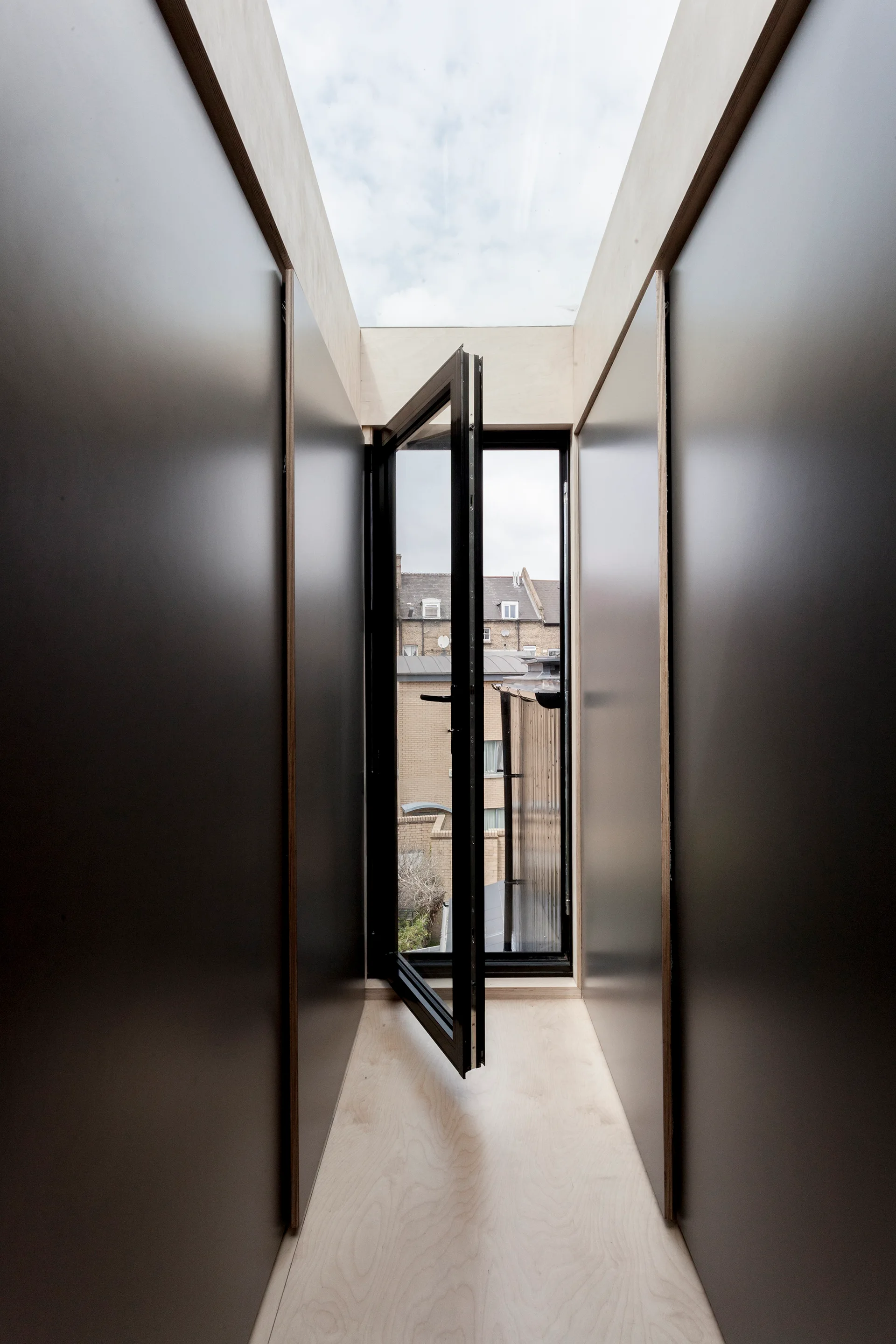 A narrow hallway features dark gray wall panels, light plywood flooring, and an open black-framed glass door under an overhead skylight.