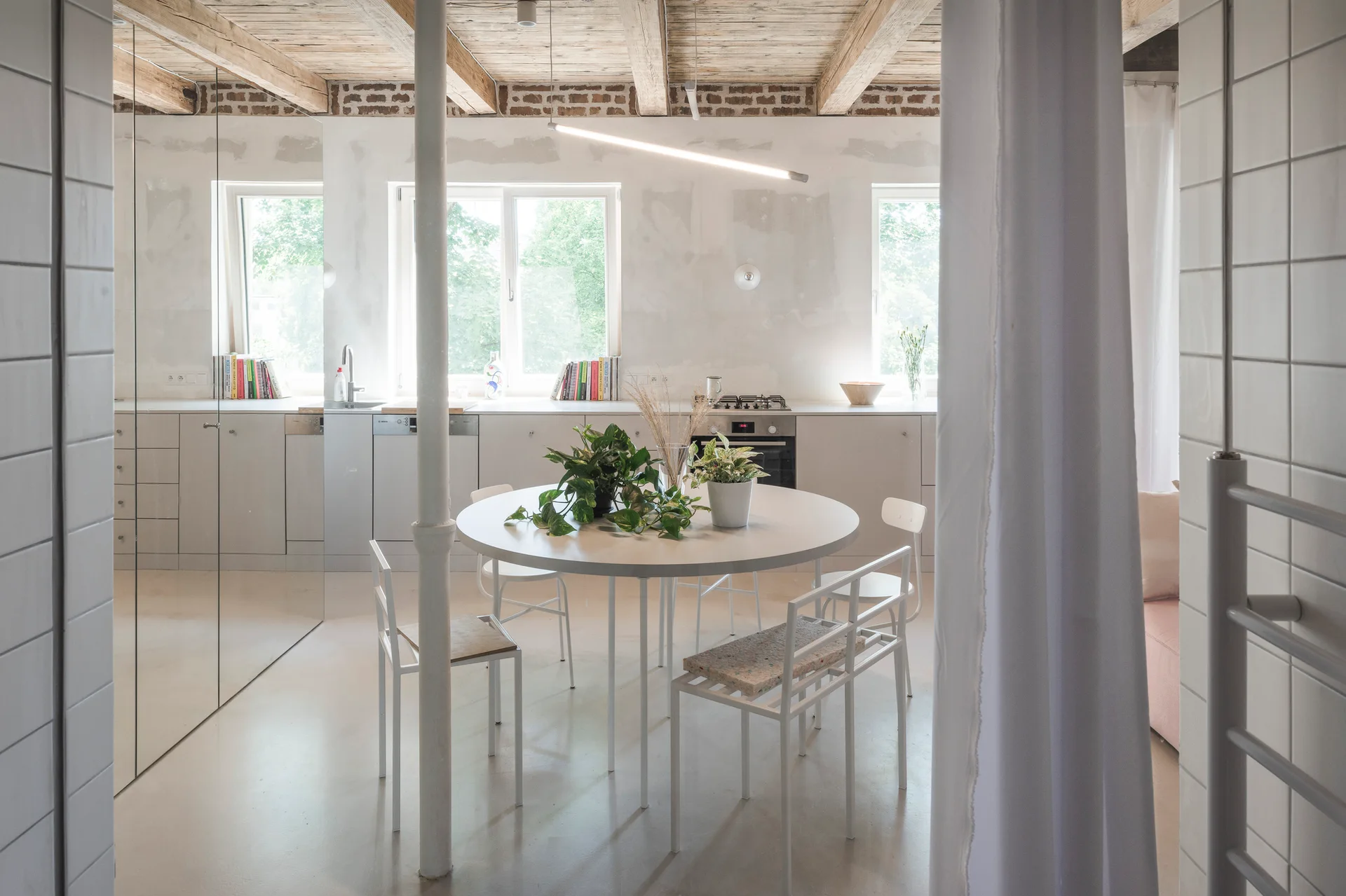A minimalist loft kitchen with exposed wood ceiling beams, raw plaster walls, light grey cabinets, and a round white dining table.