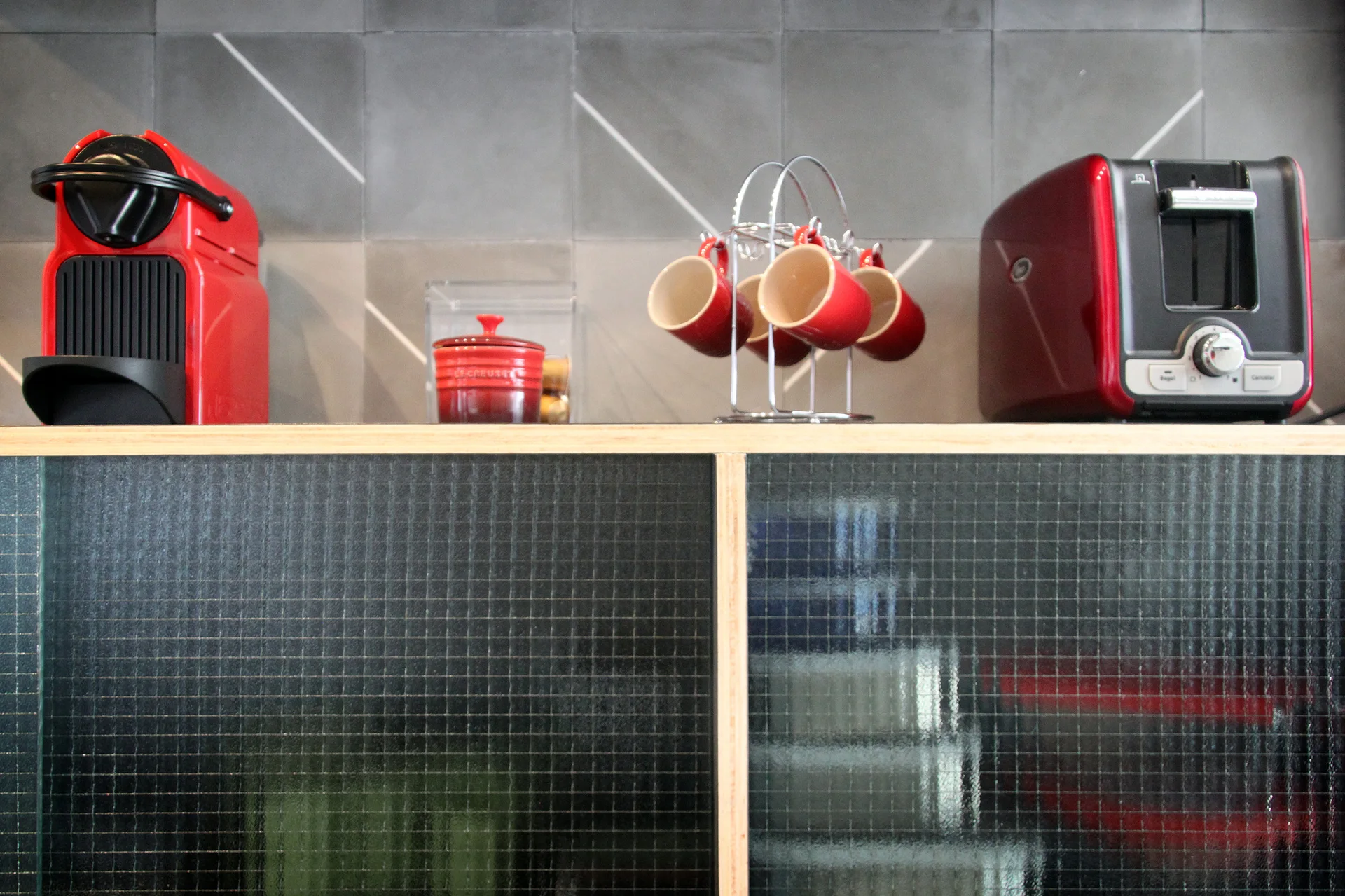 Red coffee maker, toaster, and matching mugs sit on a wooden-edged cabinet with wired glass doors against a grey tiled backsplash.