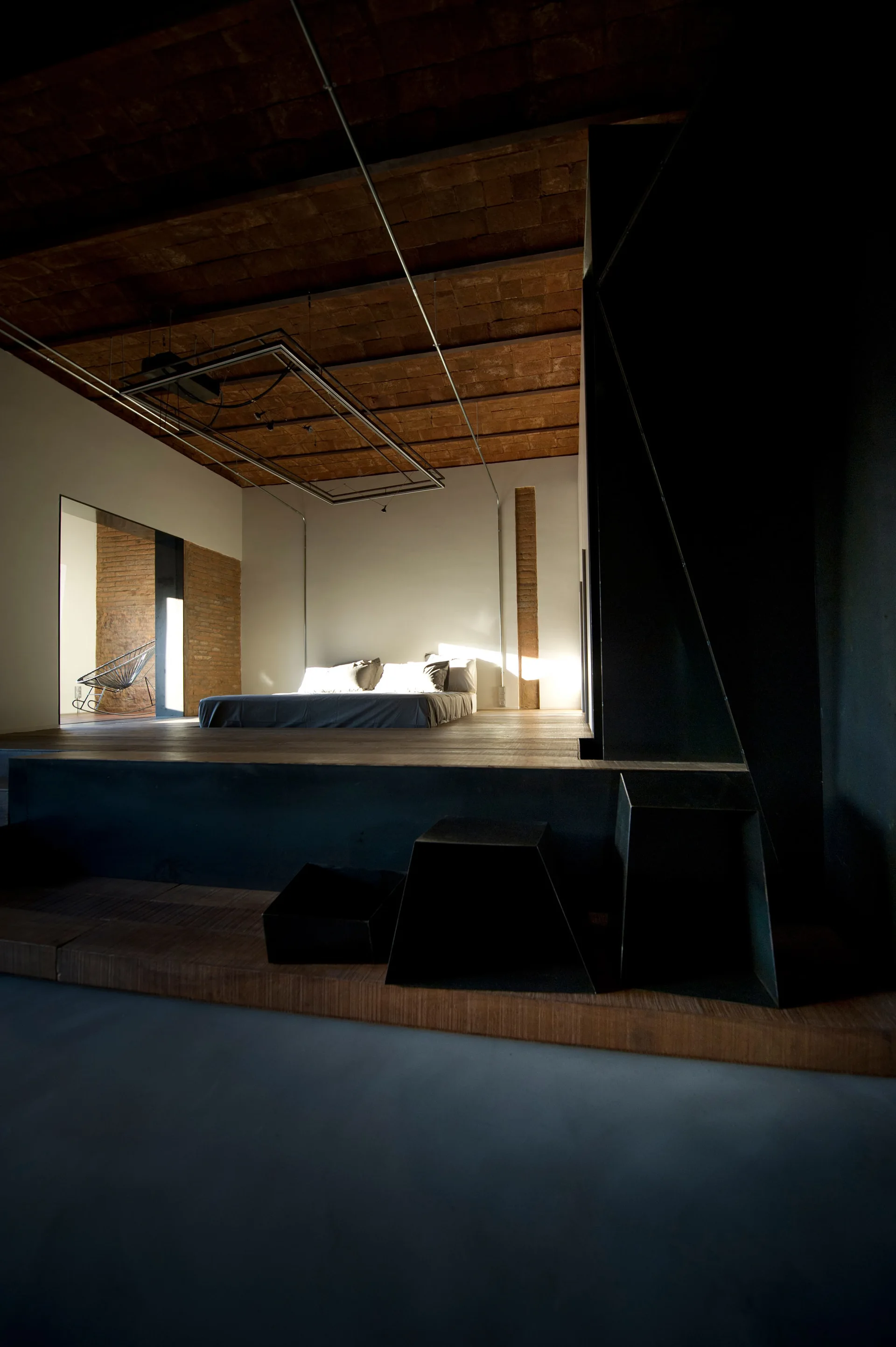 A minimalist bed rests on a raised wood platform underneath a vaulted brick ceiling in an industrial loft bedroom.
