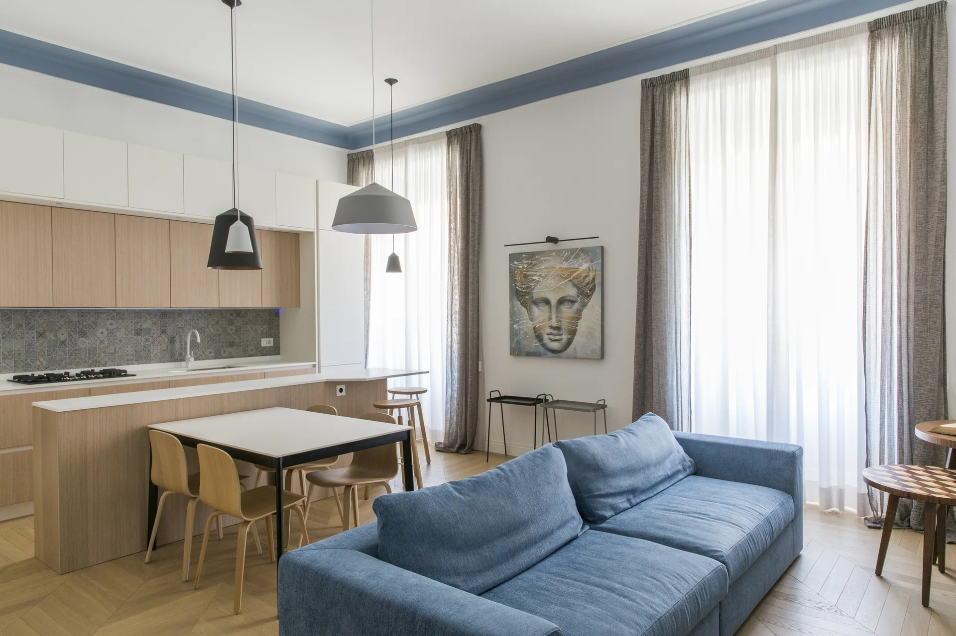 An open-concept living area features a blue sofa, chevron wood flooring, a dining table, and light wood kitchen cabinetry with a patterned backsplash.