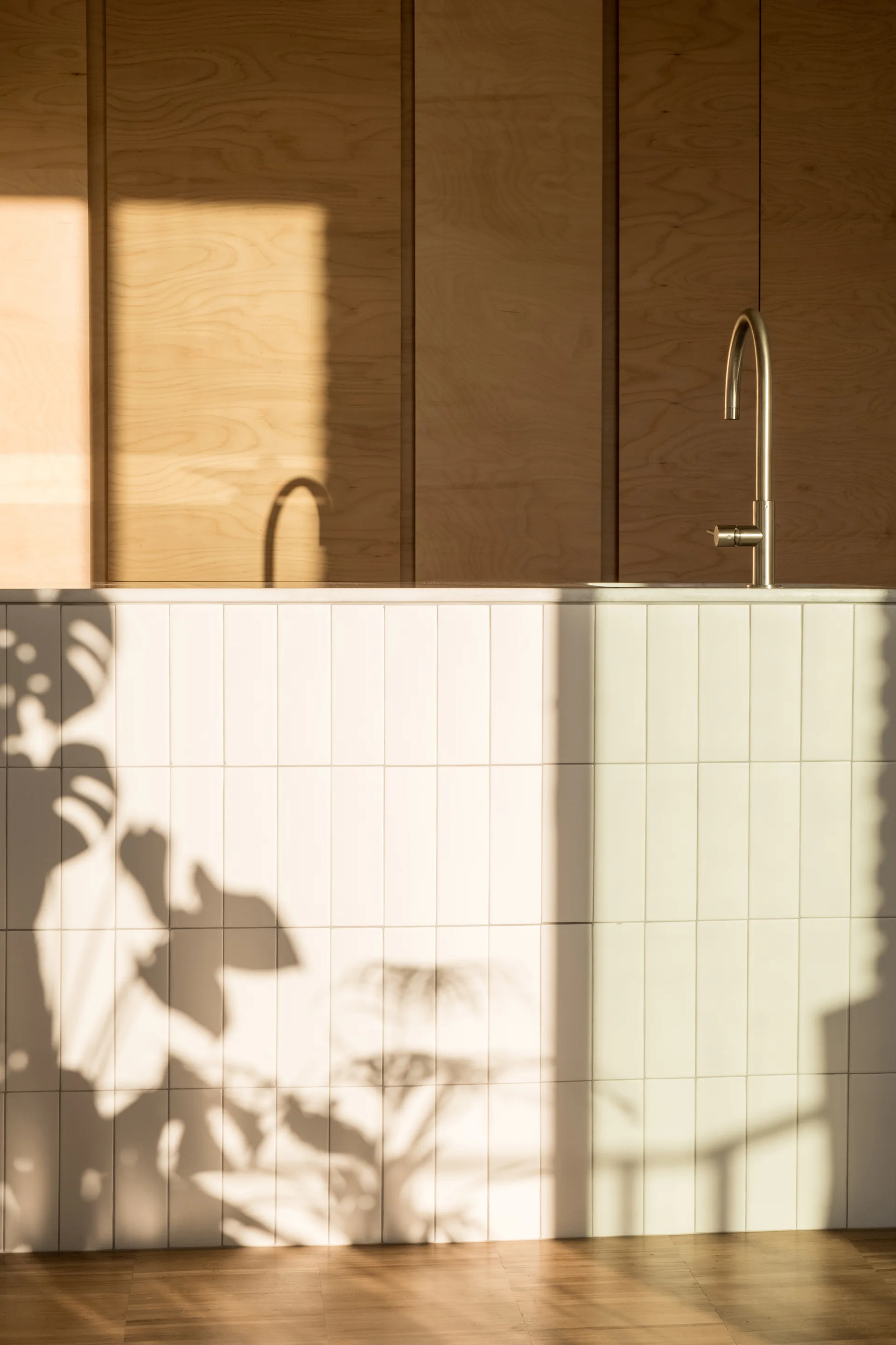 A kitchen island wrapped in white vertical tiles sits below a curved metal faucet, backed by a warm wood panel wall with dramatic leaf shadows.