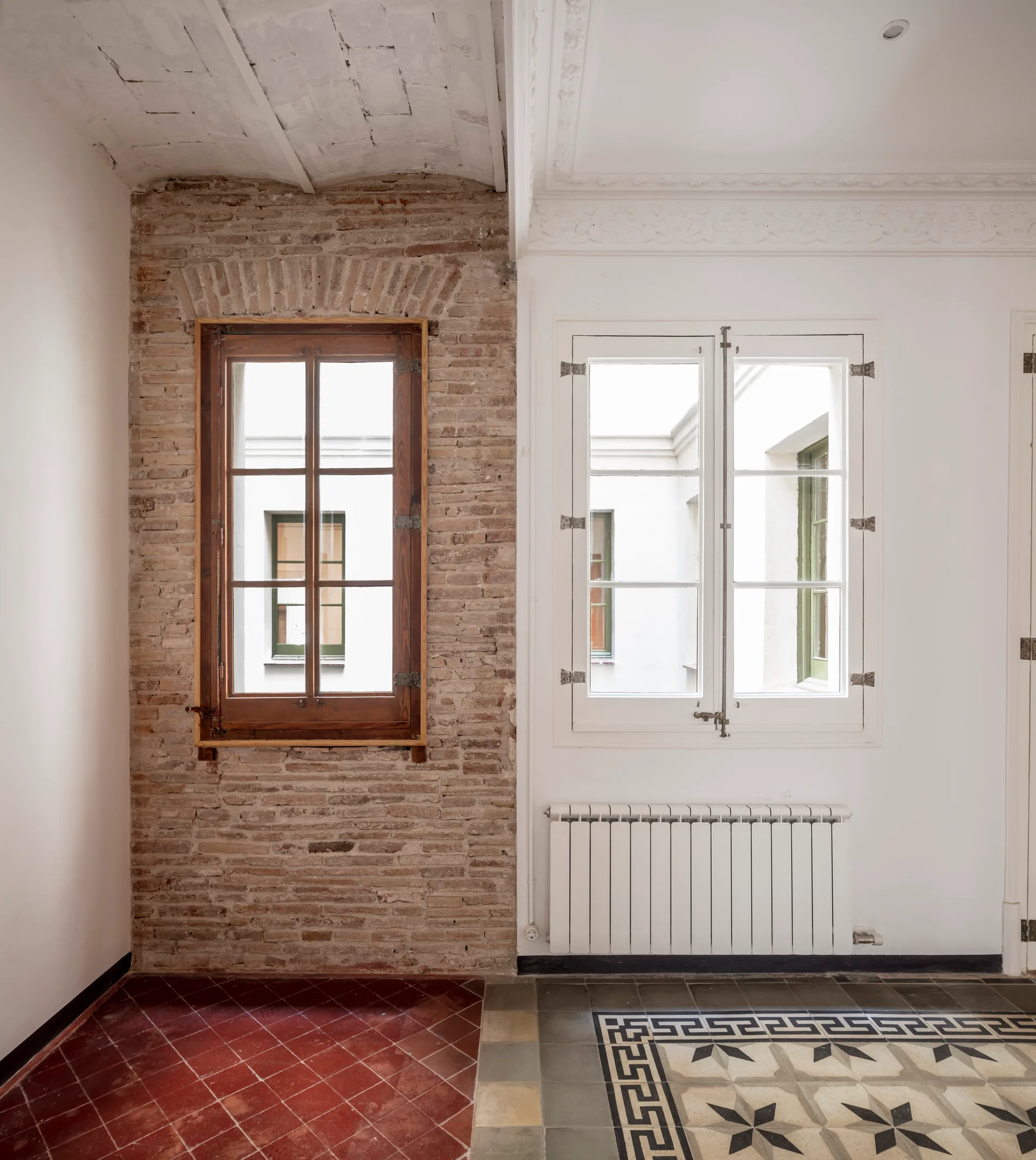 An interior space with a wall divided between exposed brick and white plaster, featuring two windows and contrasting tiled flooring.