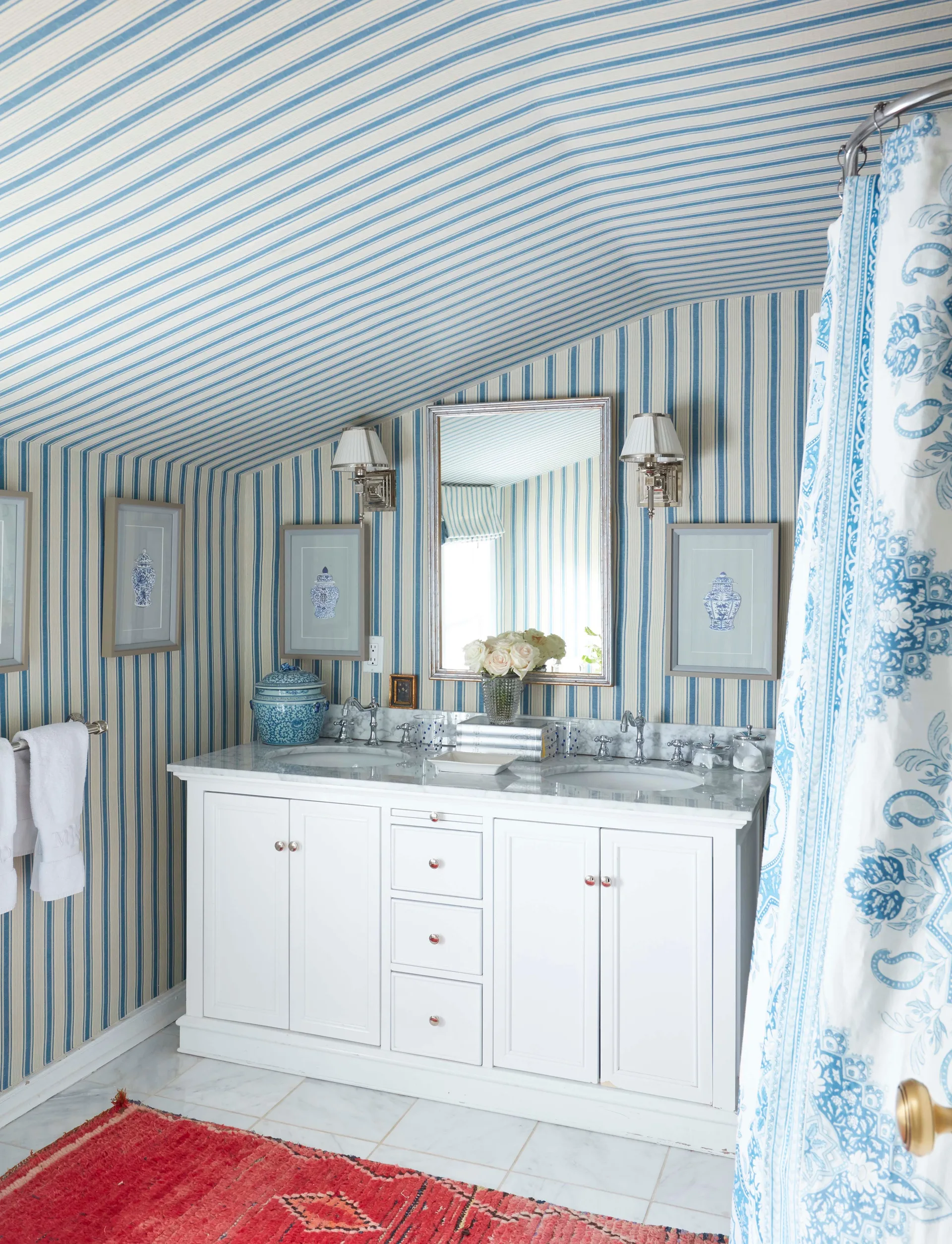 A white double vanity with a marble countertop in a bathroom featuring blue and white striped wallpaper on the walls and sloped ceiling.