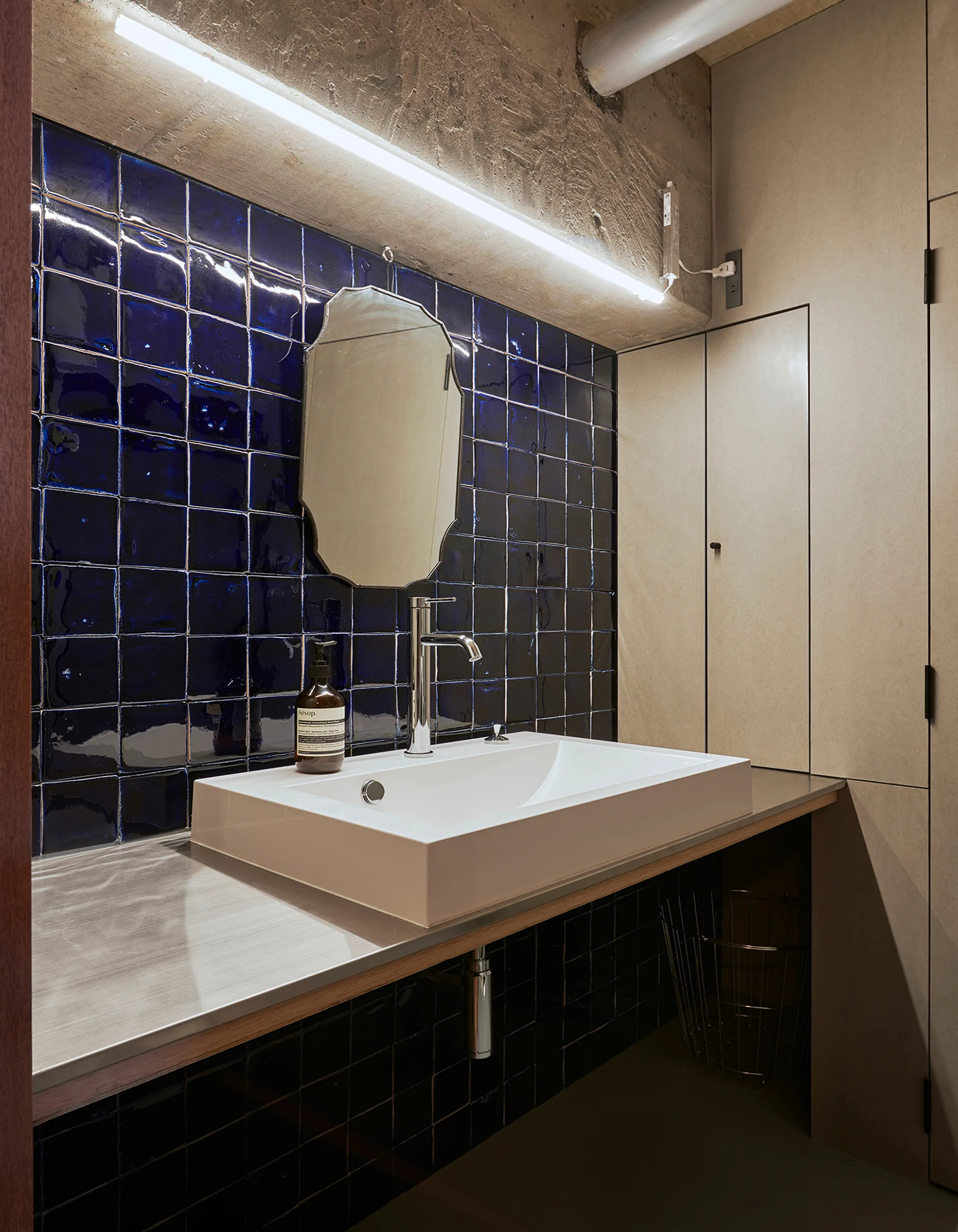 A modern bathroom featuring a white rectangular sink on a metal vanity against a glossy dark blue tiled wall beneath an exposed concrete ceiling.