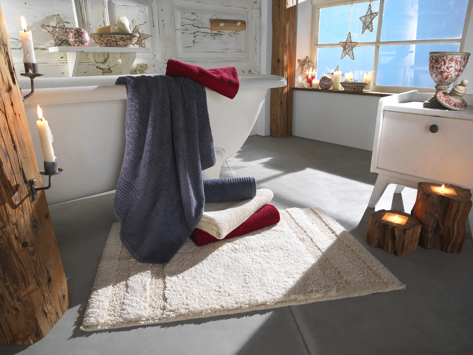 A rustic bathroom features a white bathtub with draped gray and red towels, a textured bath mat, and lit candles sitting on rough wood logs.