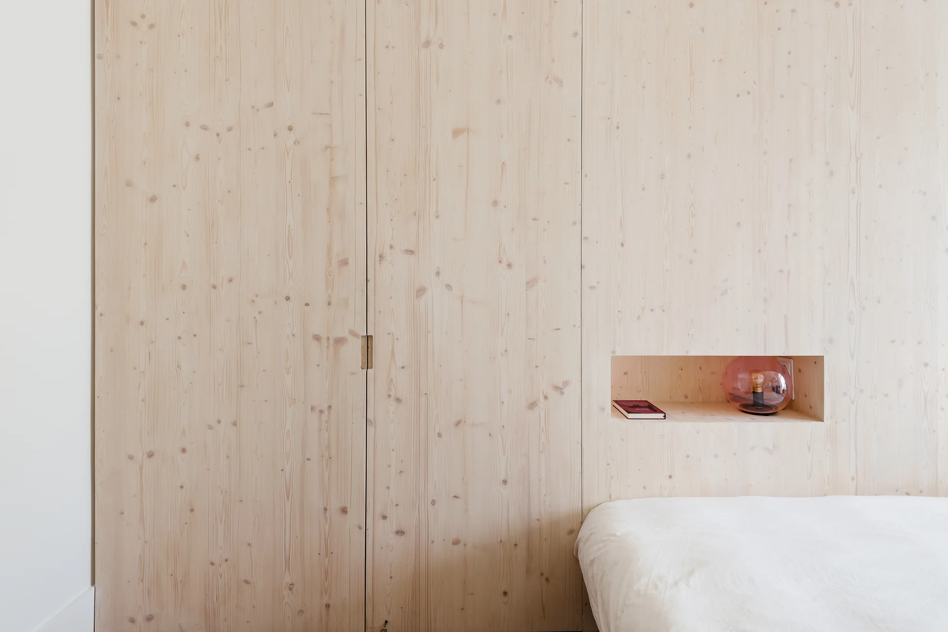 A light knotty wood paneled bedroom wall featuring integrated closet doors, a recessed shelf holding a small book and colored glass lamp, and white bedding.