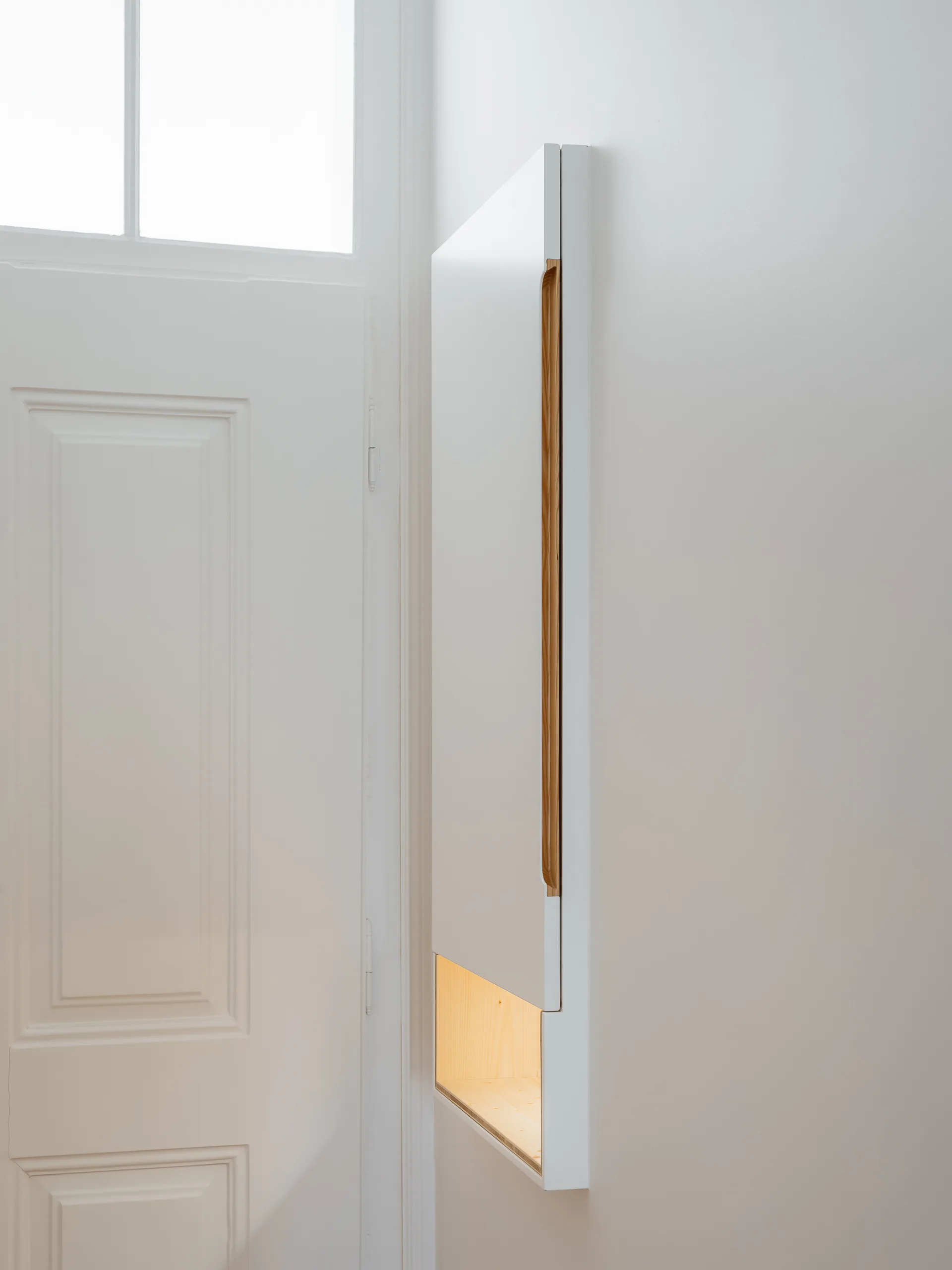 A slim white wall-mounted cabinet features a recessed wood handle and an illuminated bottom recess next to a white door.