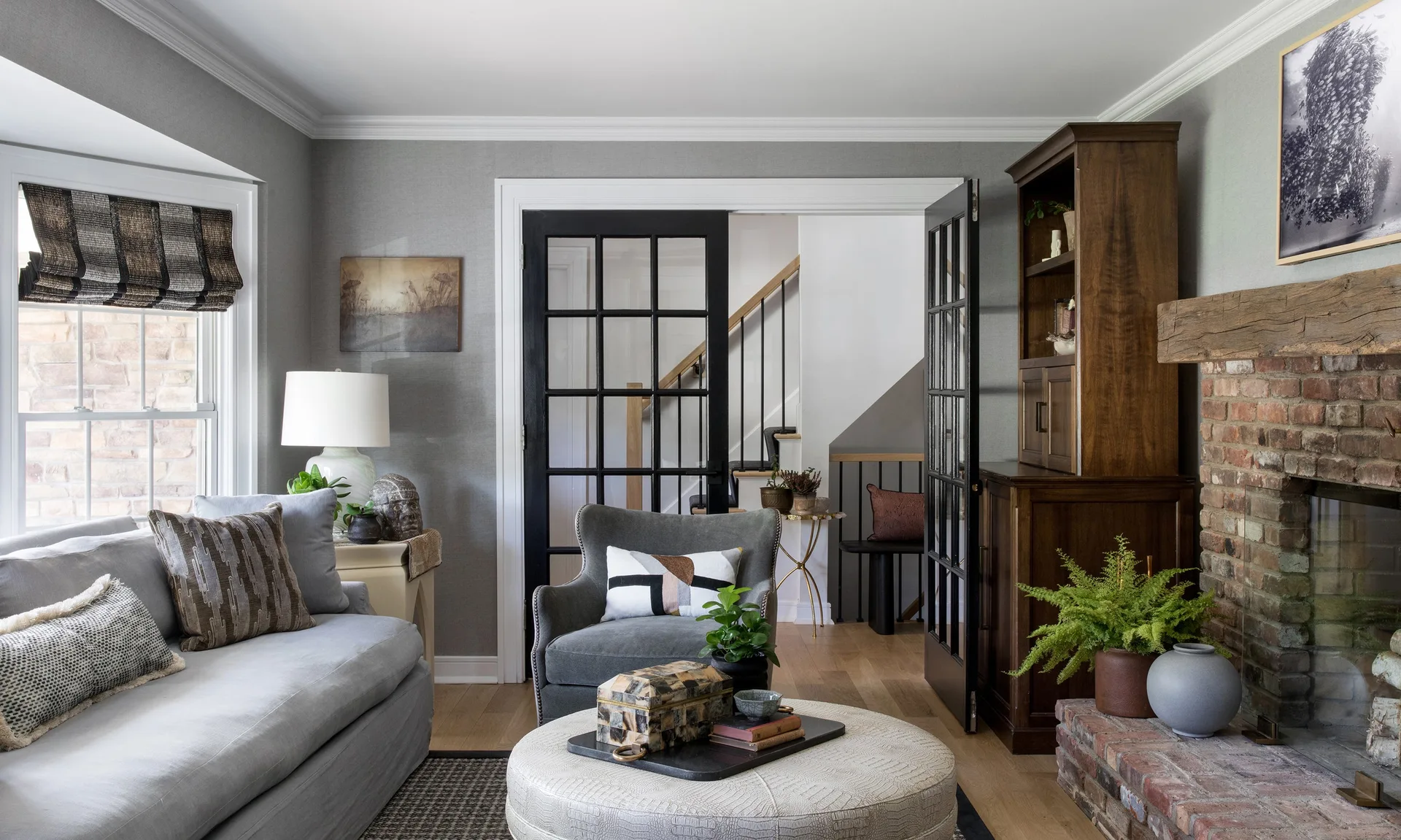 A transitional living room features a grey sofa, matching armchair, a round textured ottoman, a rustic brick fireplace, and black glass French doors.