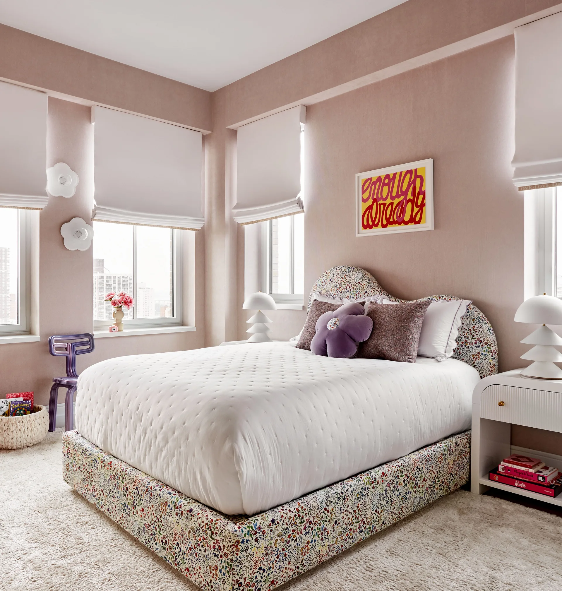 A bedroom with dusty pink walls, a floral-patterned upholstered bed, a white arched nightstand with a sculptural lamp, and white roman shades.