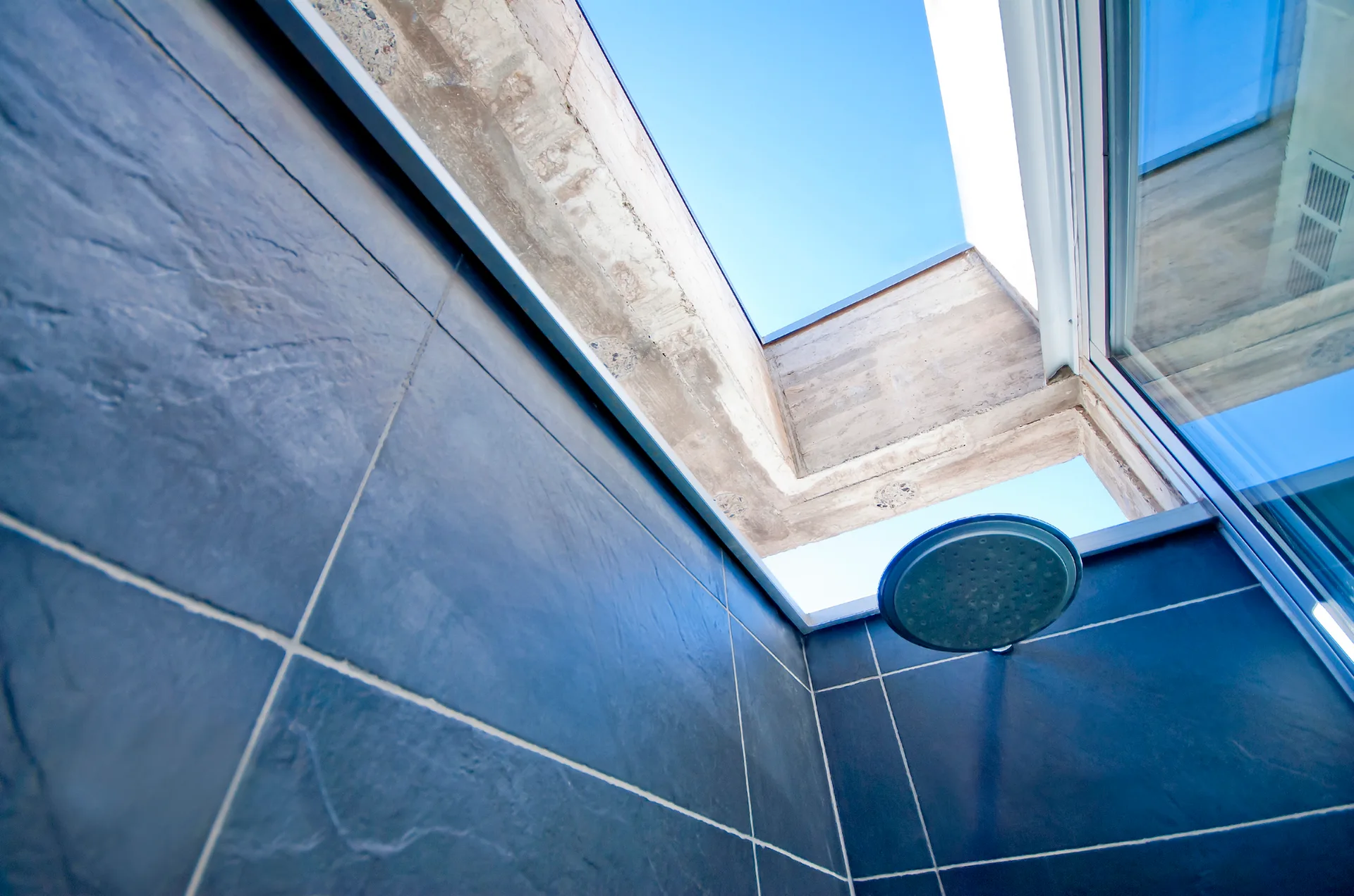 An upward angle of a shower with dark grey slate tiles, a round metal showerhead, and a rectangular skylight showing a blue sky.