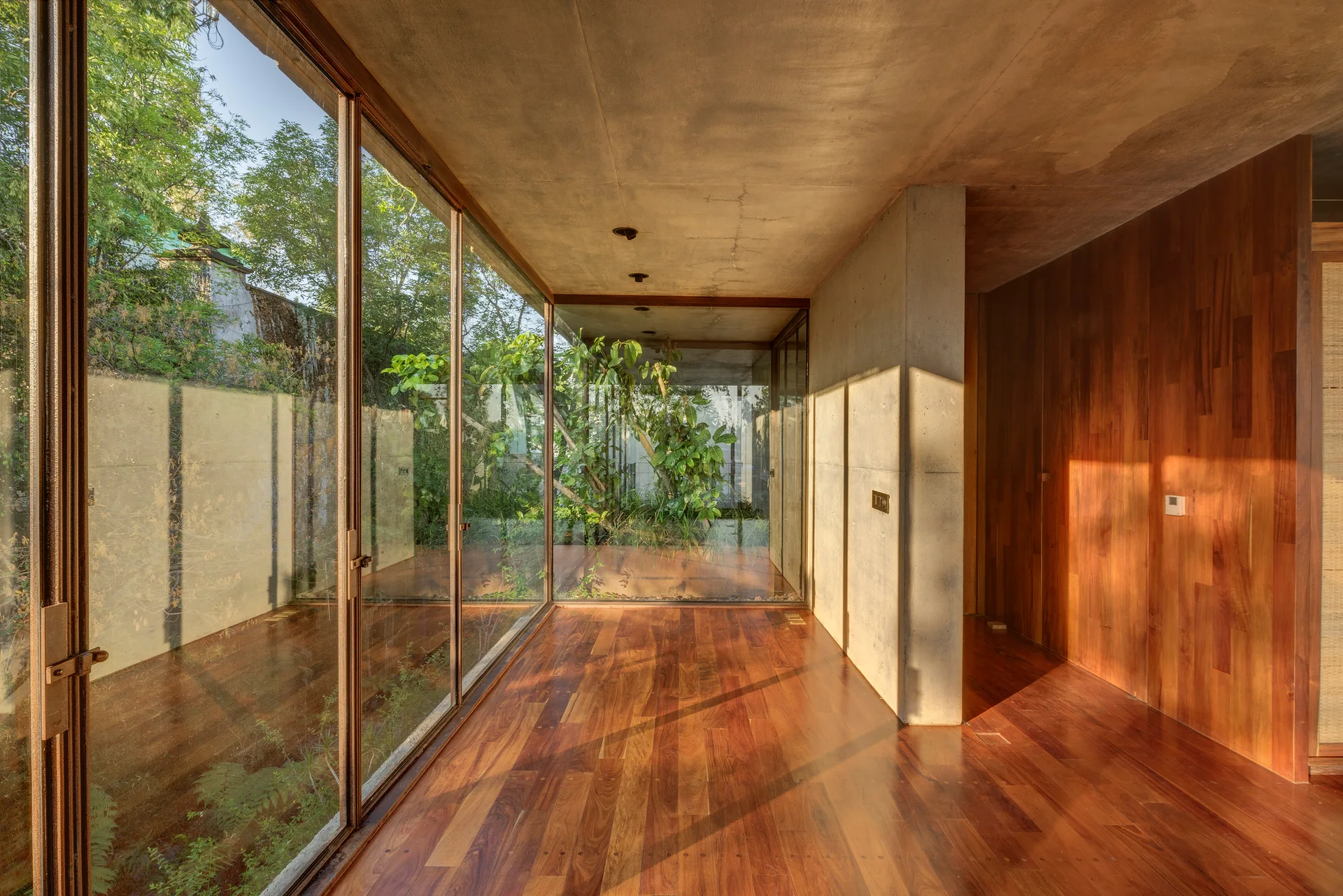 A modernist hallway features a polished wood floor, raw concrete walls and ceiling, and a floor-to-ceiling glass wall looking out to lush green trees.