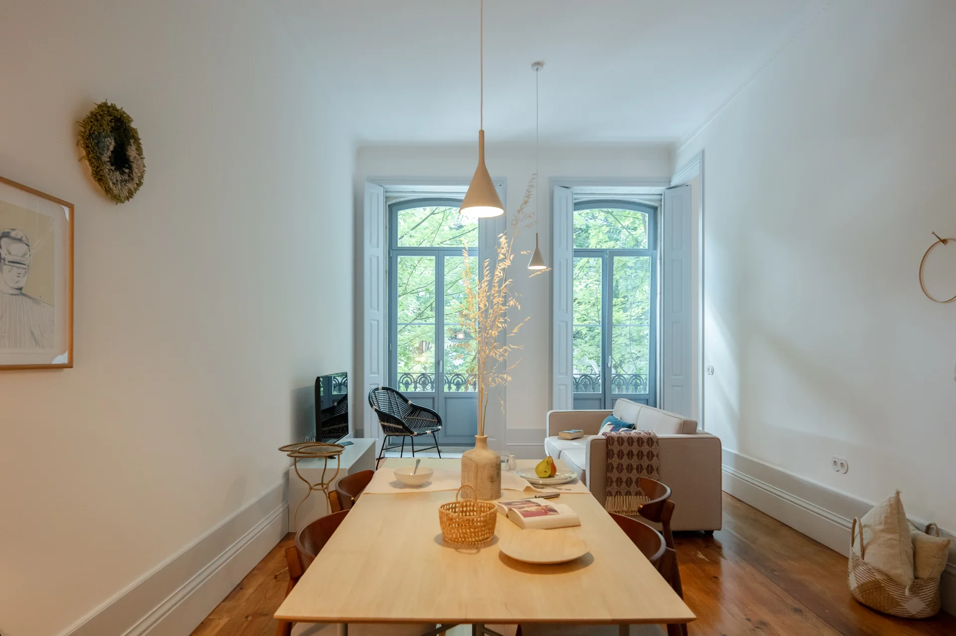 An open-plan living and dining area features a light wood dining table, white walls, and tall windows framed by pale blue interior shutters.