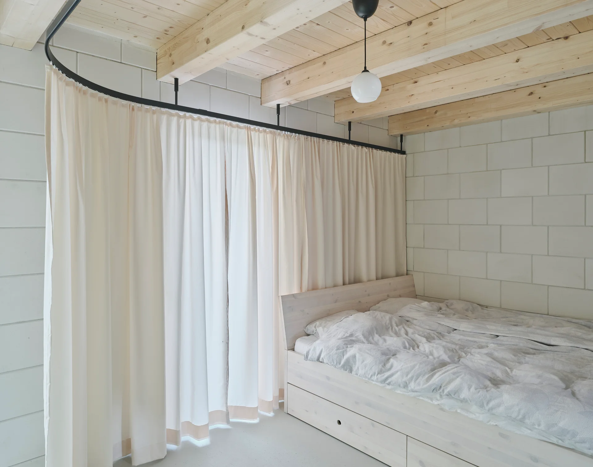 A minimalist bedroom features a light wood platform bed against white concrete block walls under an exposed timber ceiling with a curved curtain rail.