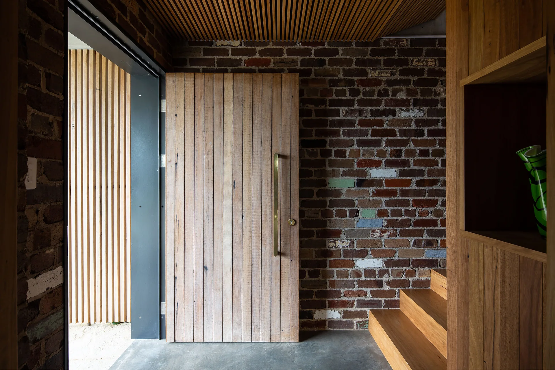 A residential entryway featuring an exposed brick wall, a vertical-slat wooden door with a brass handle, concrete flooring, and wooden stairs.