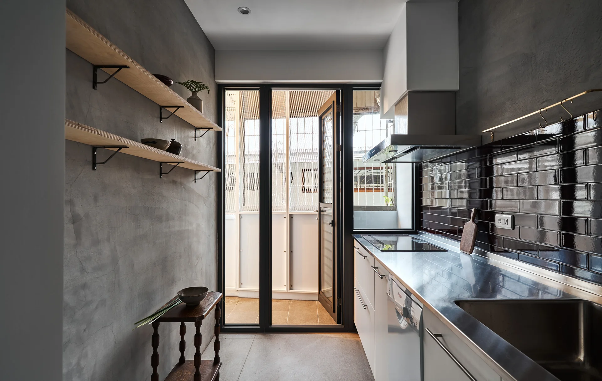 A narrow galley kitchen features textured concrete walls, wooden floating shelves, white cabinets under stainless steel countertops, and dark glossy tile.