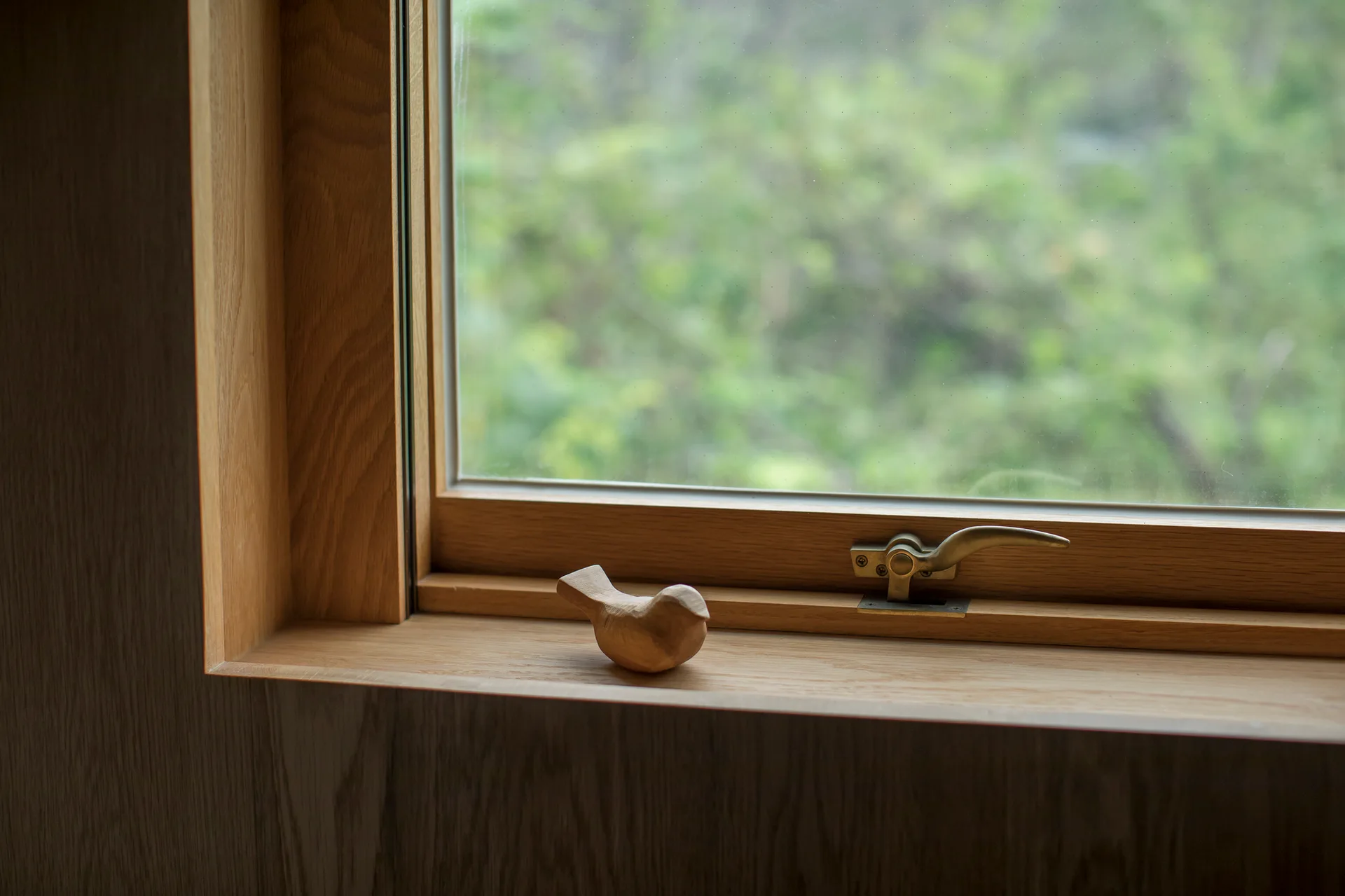 A small carved wooden bird figurine sits on a deep natural wood window sill next to a metal casement latch with green foliage visible outside.
