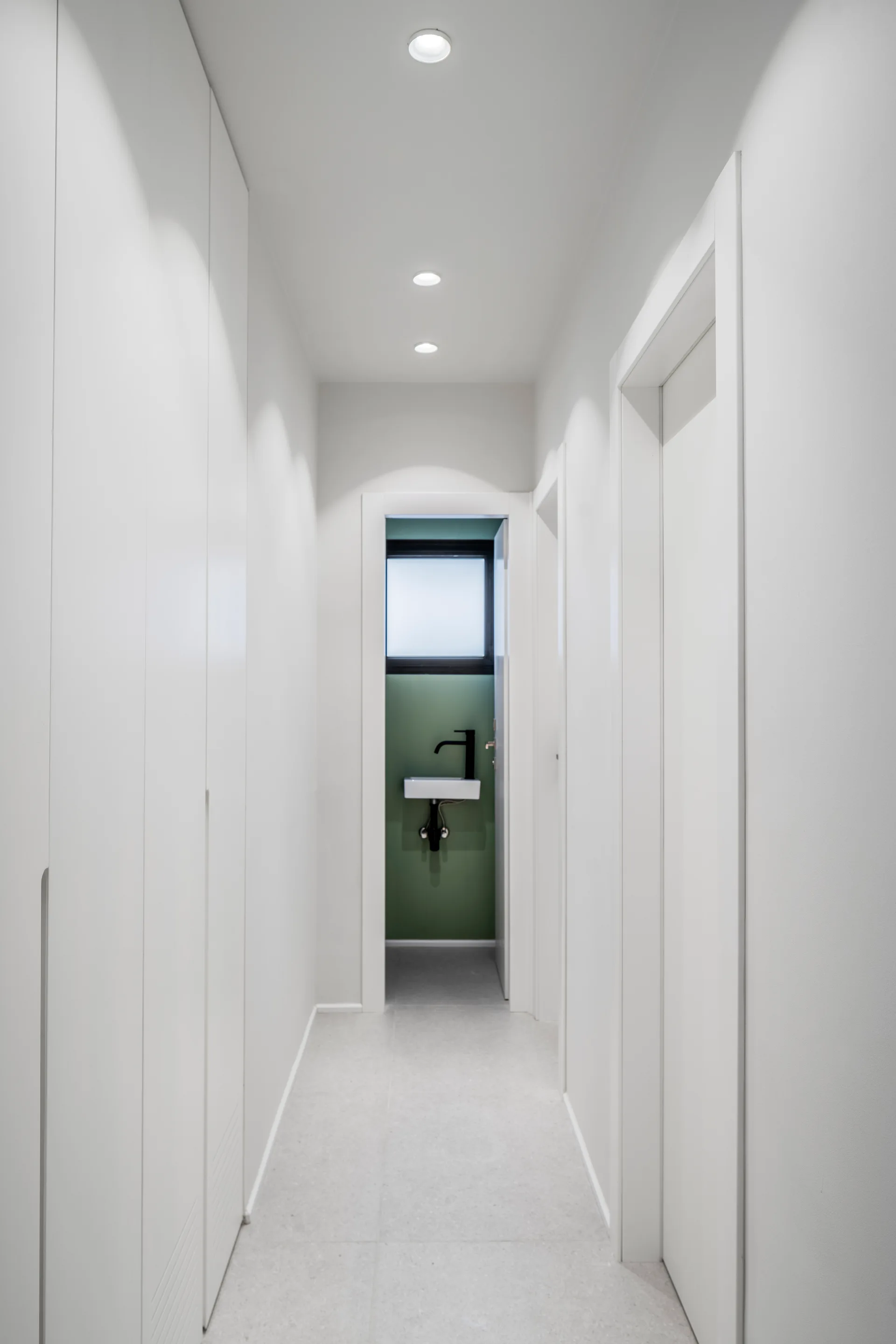 A narrow white hallway with floor-to-ceiling built-in cabinets leads to an open doorway showing a small bathroom with a green wall and tiny sink.