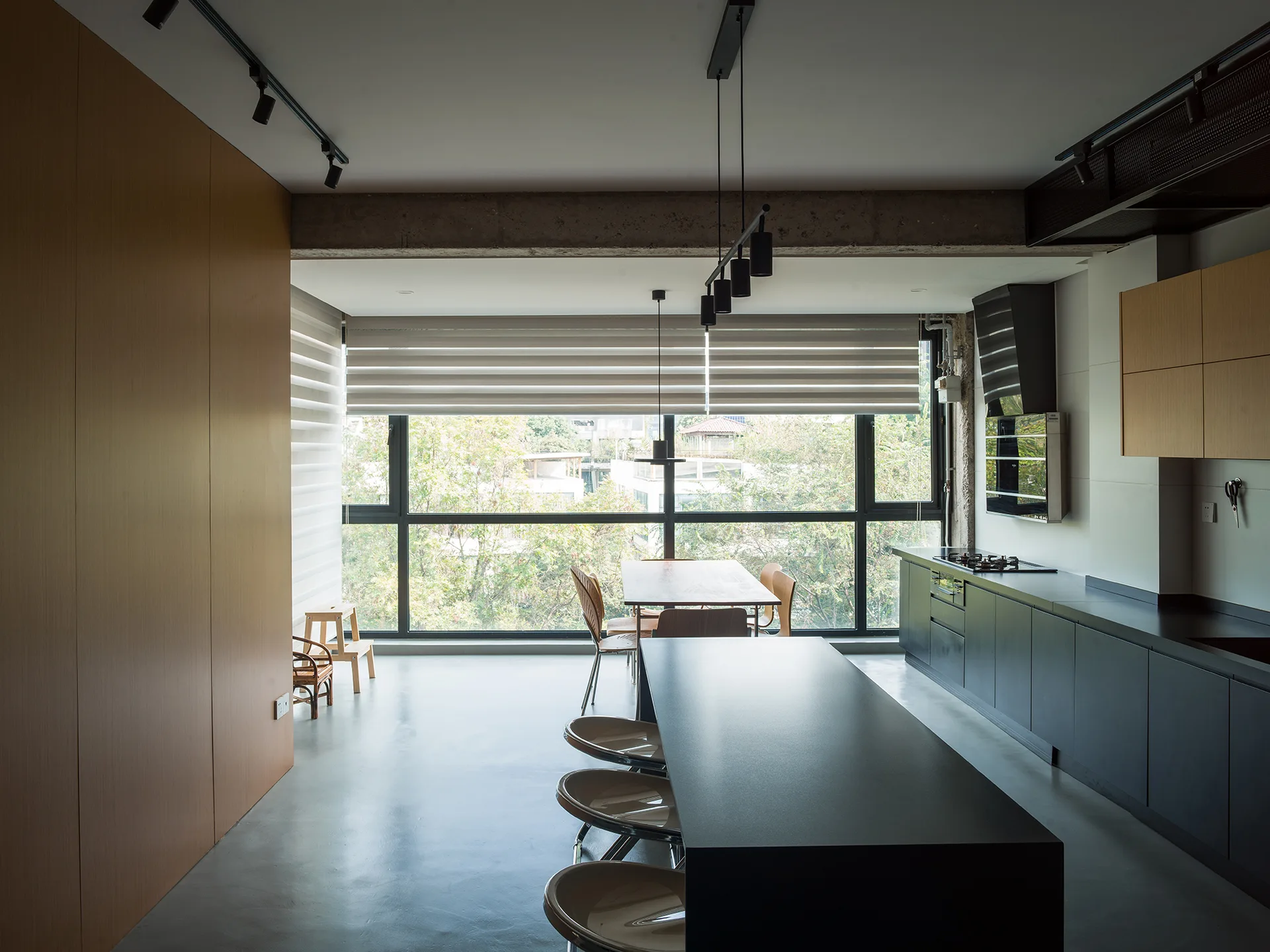 A modern industrial kitchen features a long black island with stools, dark cabinetry, an exposed concrete ceiling beam, and a large window.