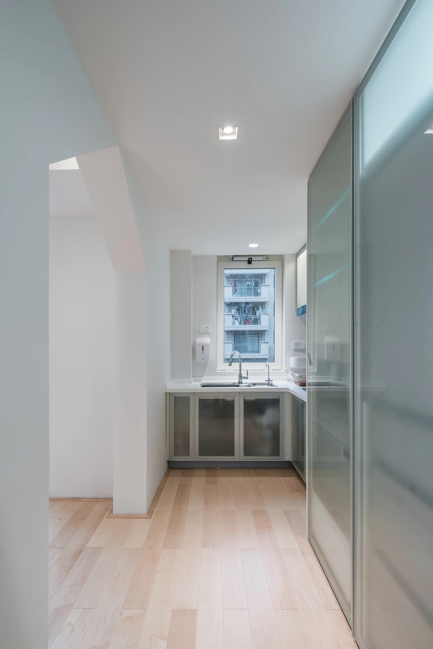 A minimalist kitchen features light wood flooring, a stainless steel sink below a window, and large frosted glass sliding partition doors.