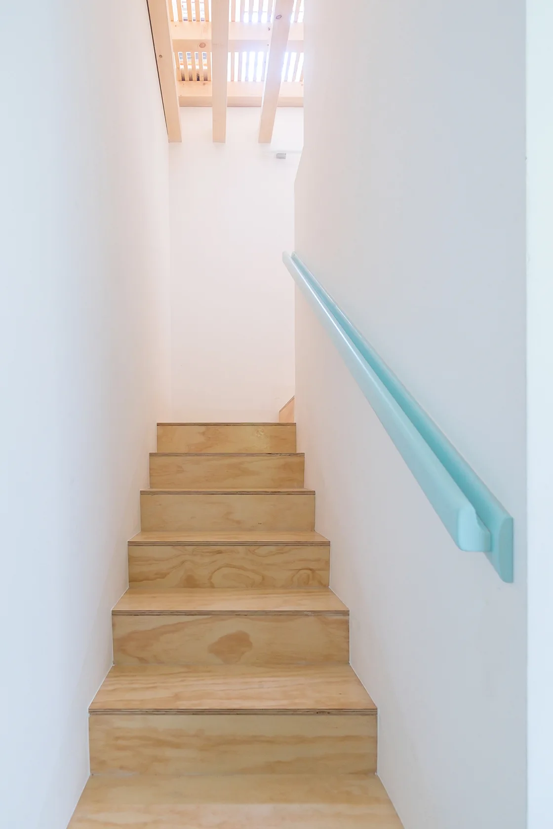 A minimalist indoor staircase featuring light wood plywood steps, clean white walls, and a light blue wall-mounted handrail.