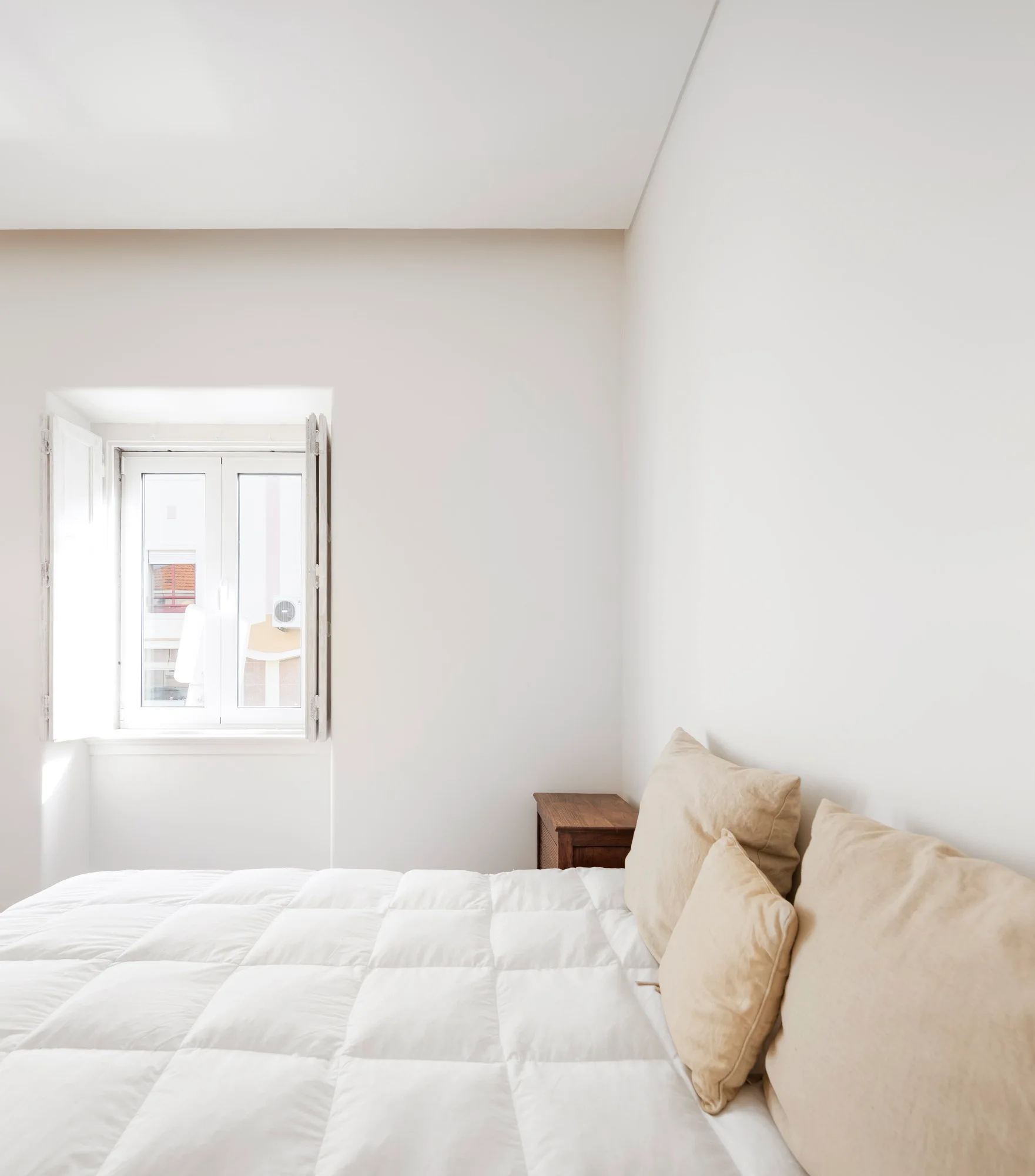 A minimalist white bedroom features a bed with a white quilted duvet and beige pillows next to a recessed window with open white shutters.