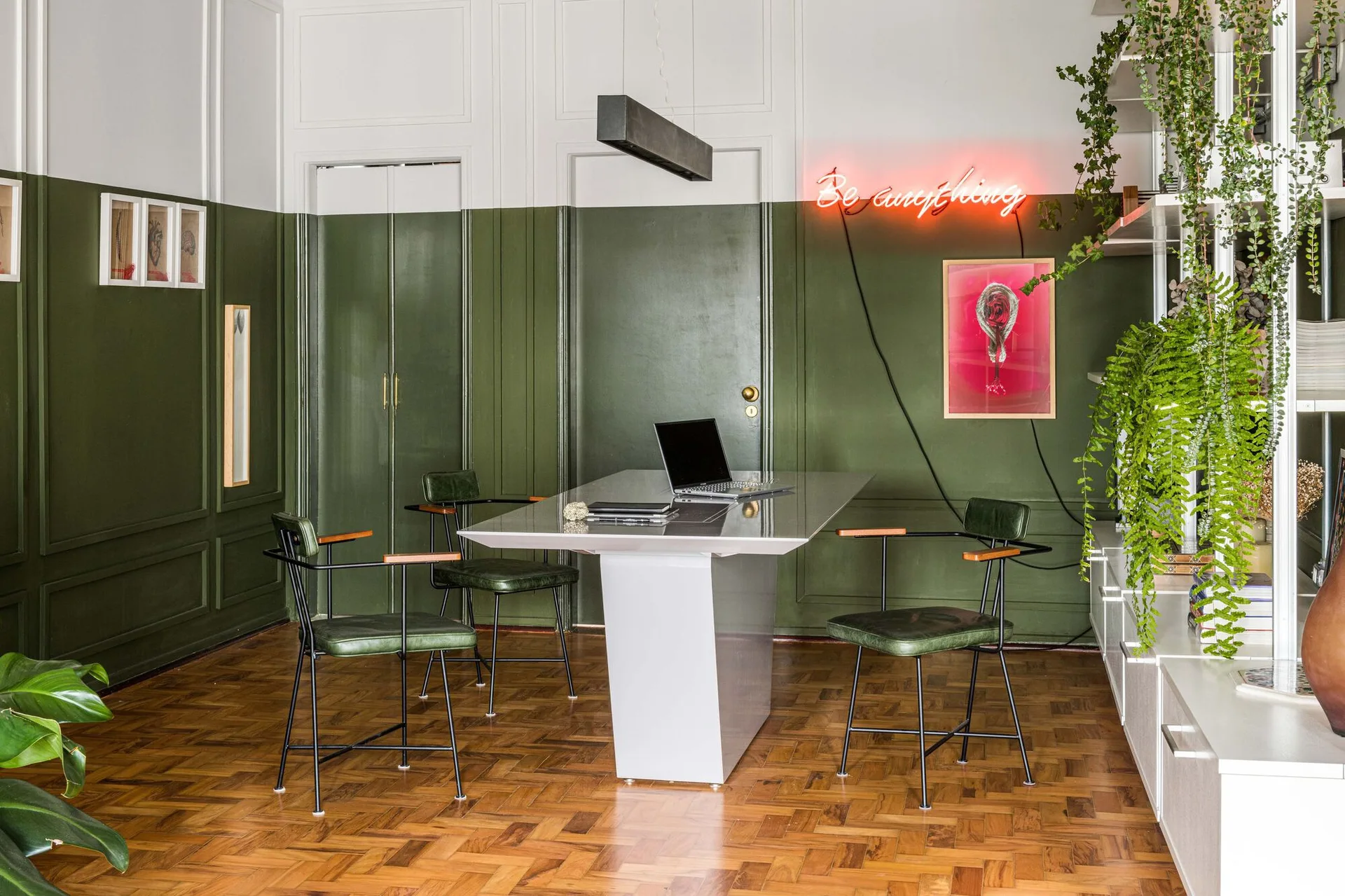 A home office with olive green wall paneling, a modern white desk, matching green leather chairs, parquet wood flooring, and a glowing neon sign.