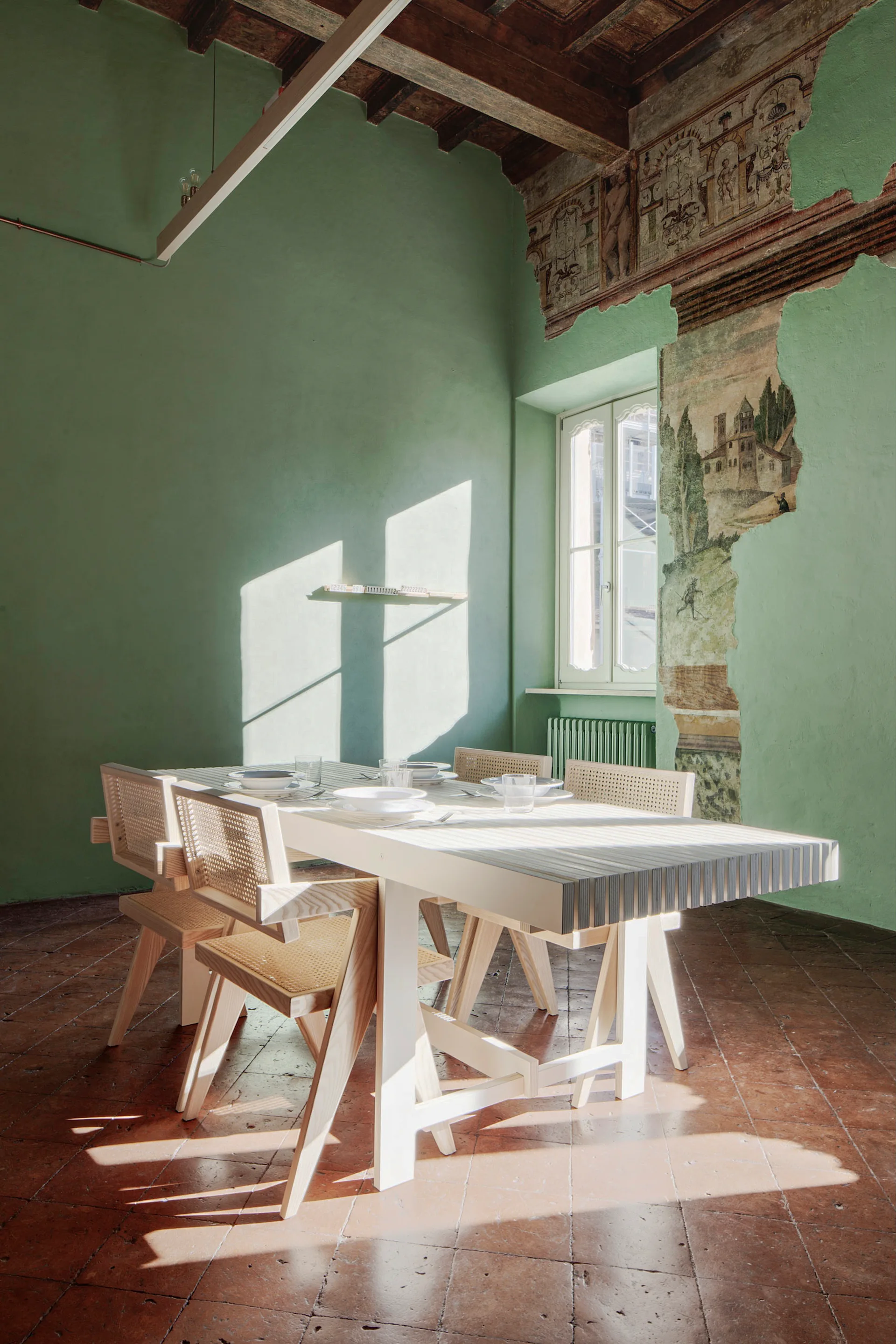A white wooden dining table and cane chairs sit on a terracotta tiled floor in a room with pale green walls, exposed ceiling beams, and a wall fresco.