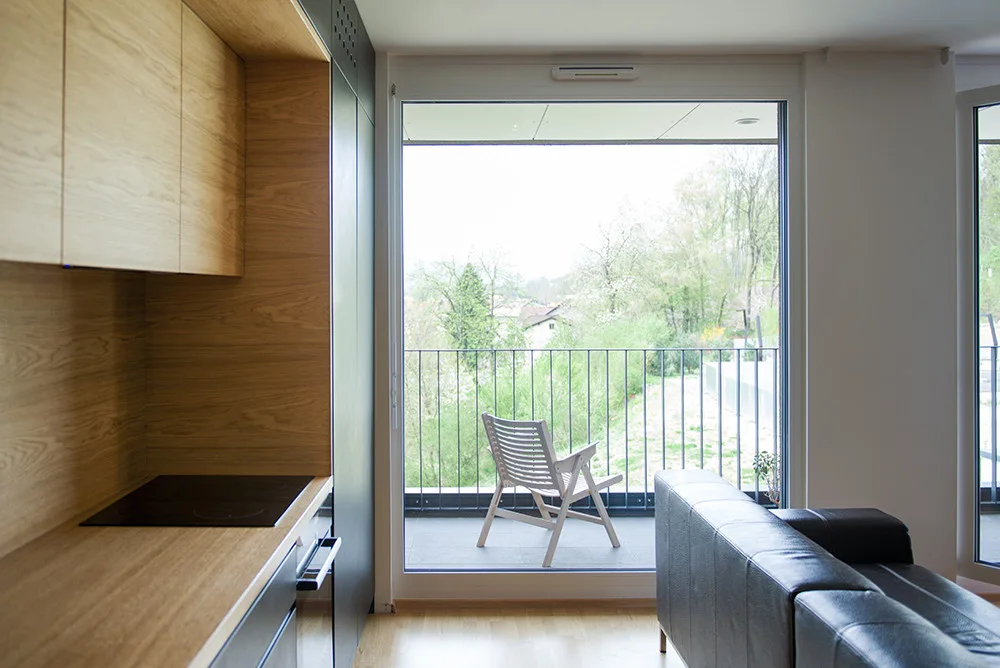 A modern interior features light wood kitchen cabinetry on the left and a dark leather sofa facing a large window that opens onto a balcony.