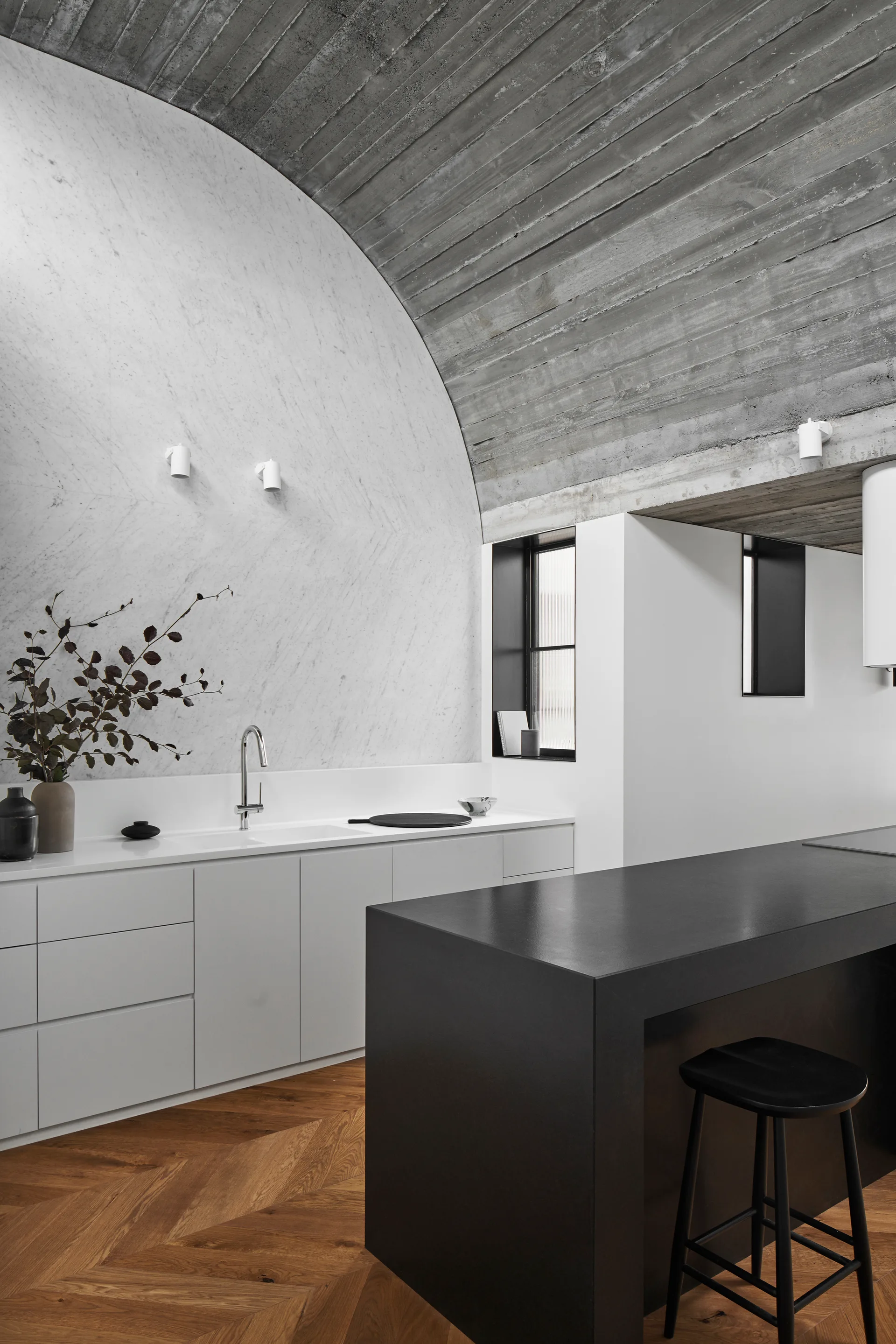 A minimalist kitchen features a gray concrete vaulted ceiling, a curved white marble wall, a dark kitchen island, and herringbone wood flooring.