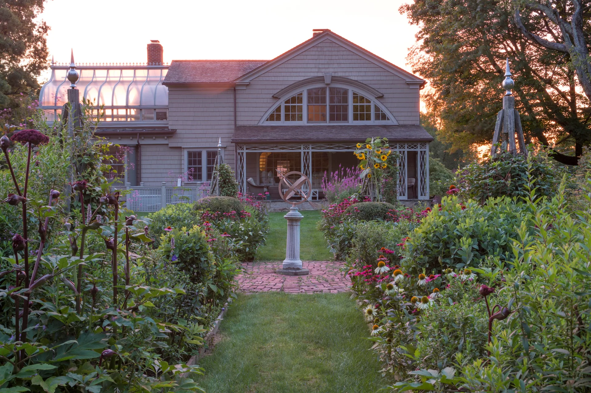 A brick path winds through a lush flower garden toward a stone pedestal sundial in front of a shingled house with a glass conservatory.