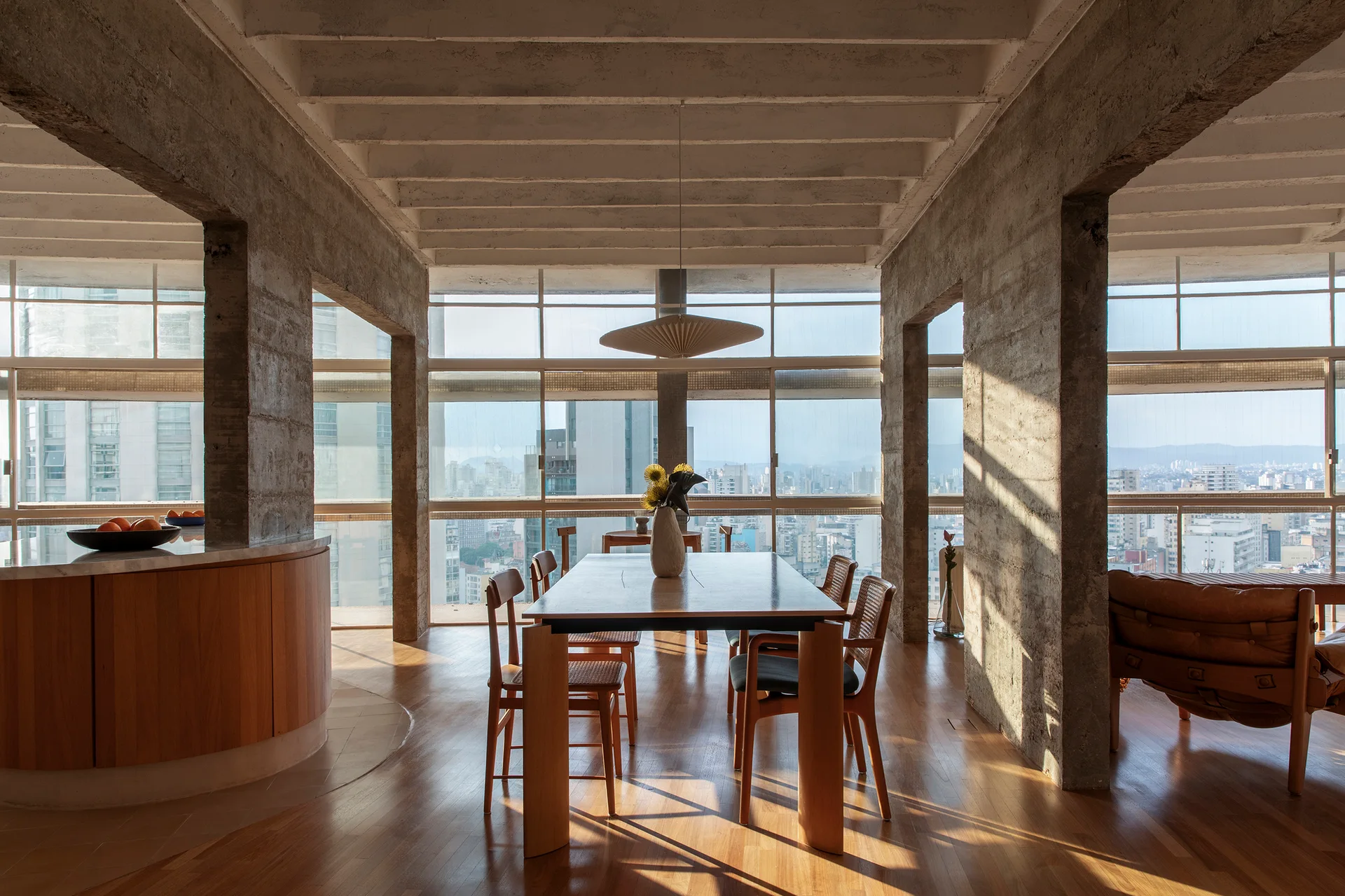 A sunlit dining room features a wooden table and chairs between massive concrete columns under an exposed waffle slab ceiling with city views.