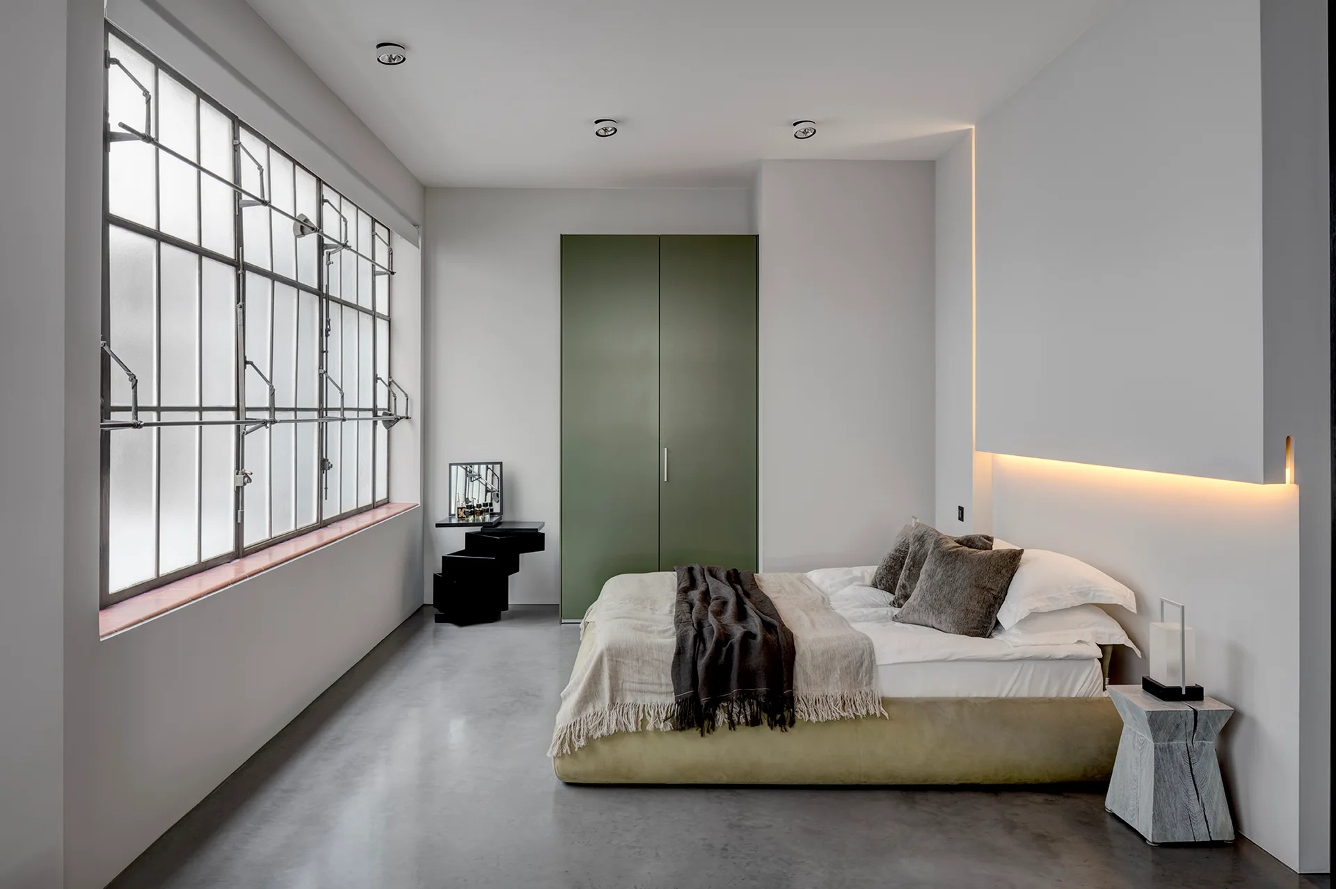 A minimalist bedroom featuring a low-profile bed, an integrated olive green wardrobe, and a large metal-framed industrial window.