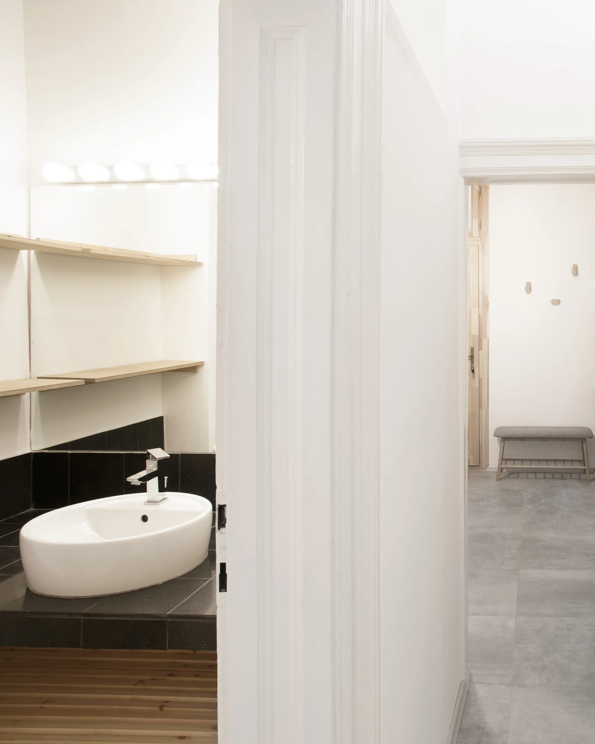 A white ceramic vessel sink rests on a black tiled vanity next to open wood shelves, viewed through a doorway opposite a gray tiled hallway.