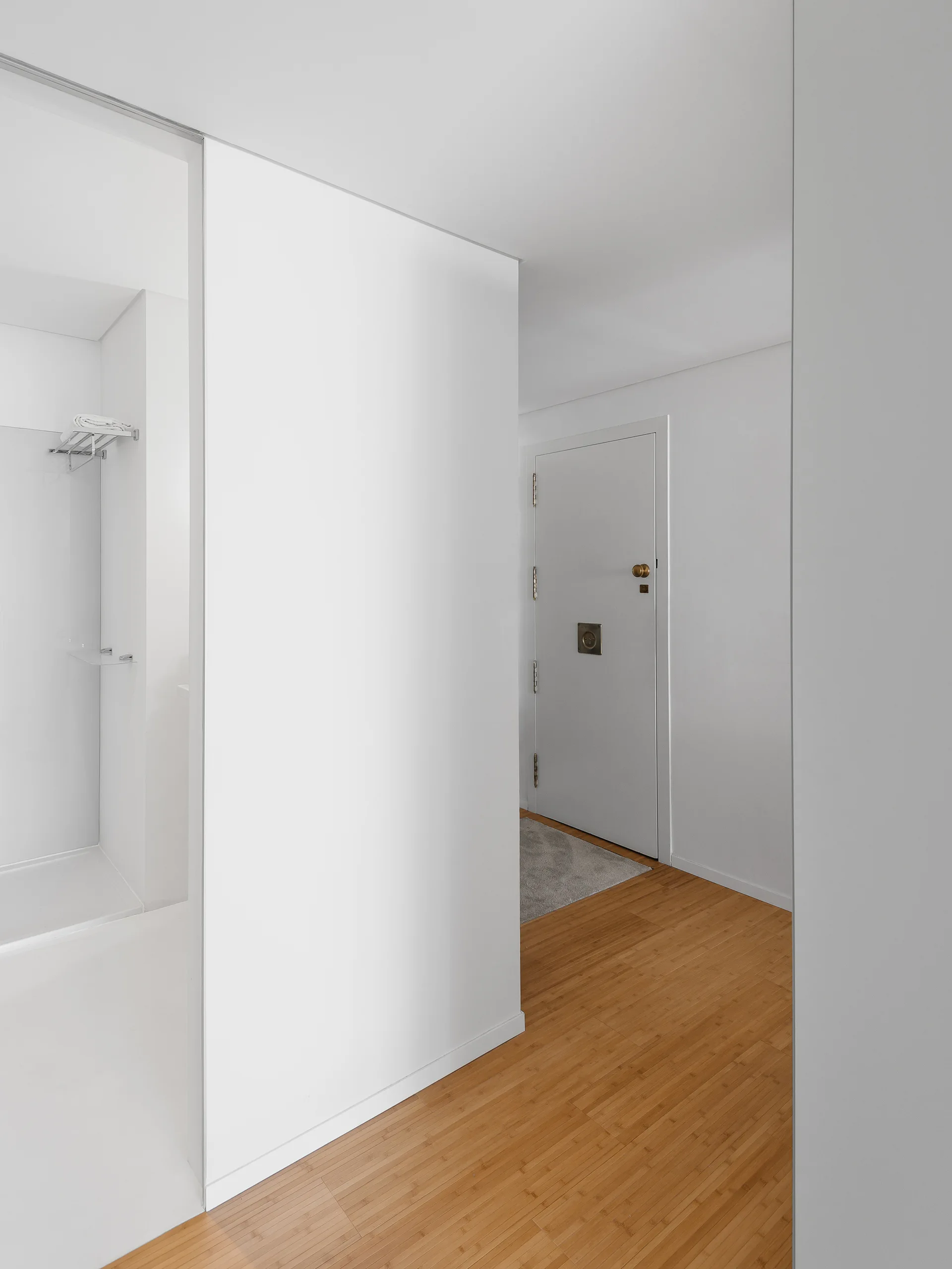 A minimalist white entryway with a wooden floor leads past a bathroom doorway toward a white entrance door with brass hardware.