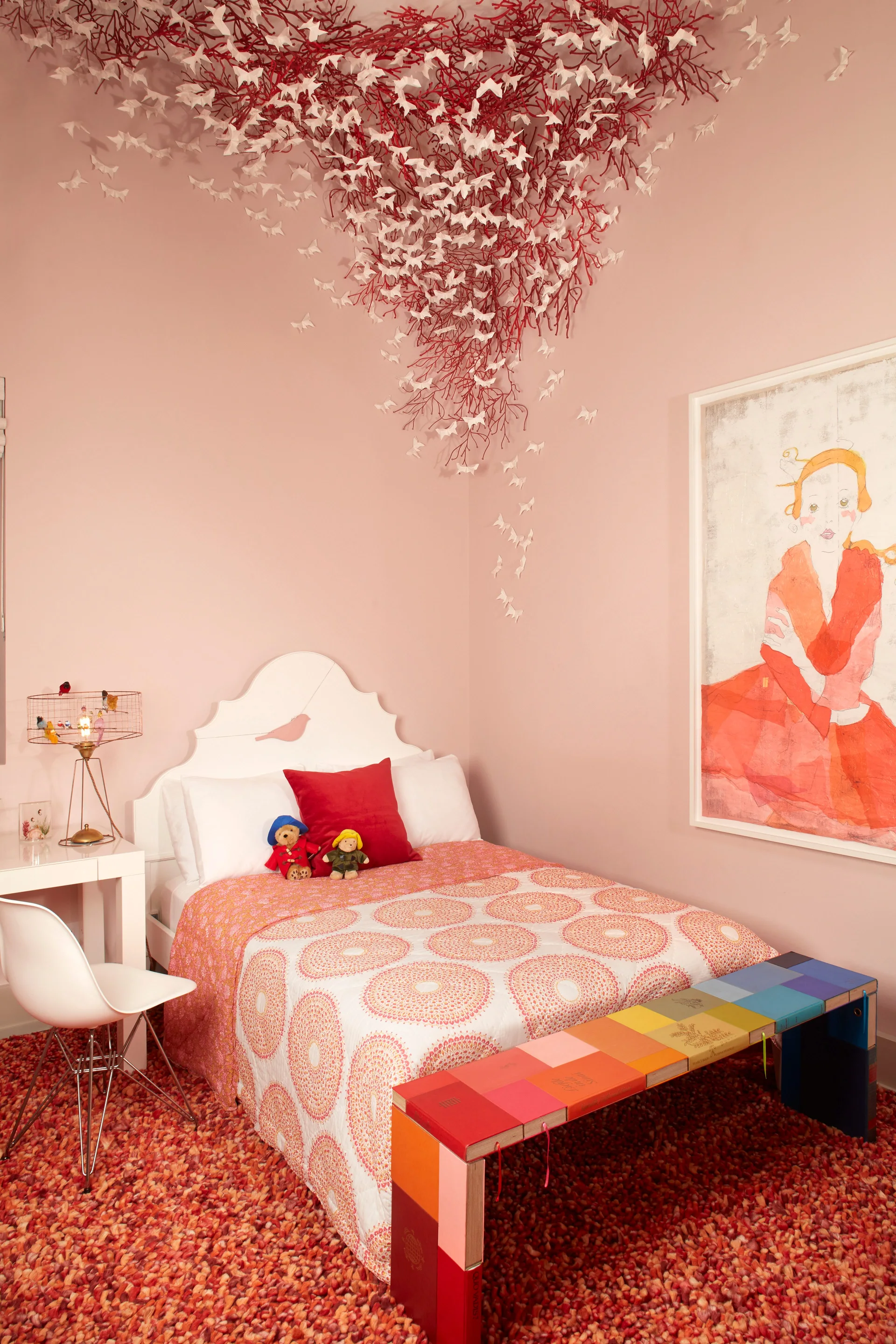 A children's bedroom features pink walls, a white bed, a colorful book bench, and a red branch ceiling installation with paper butterflies.