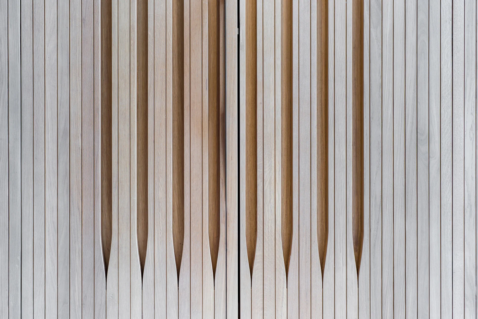 A close-up view of a light-colored wooden wall featuring vertical slats and deeply carved recessed channels that taper at the bottom.