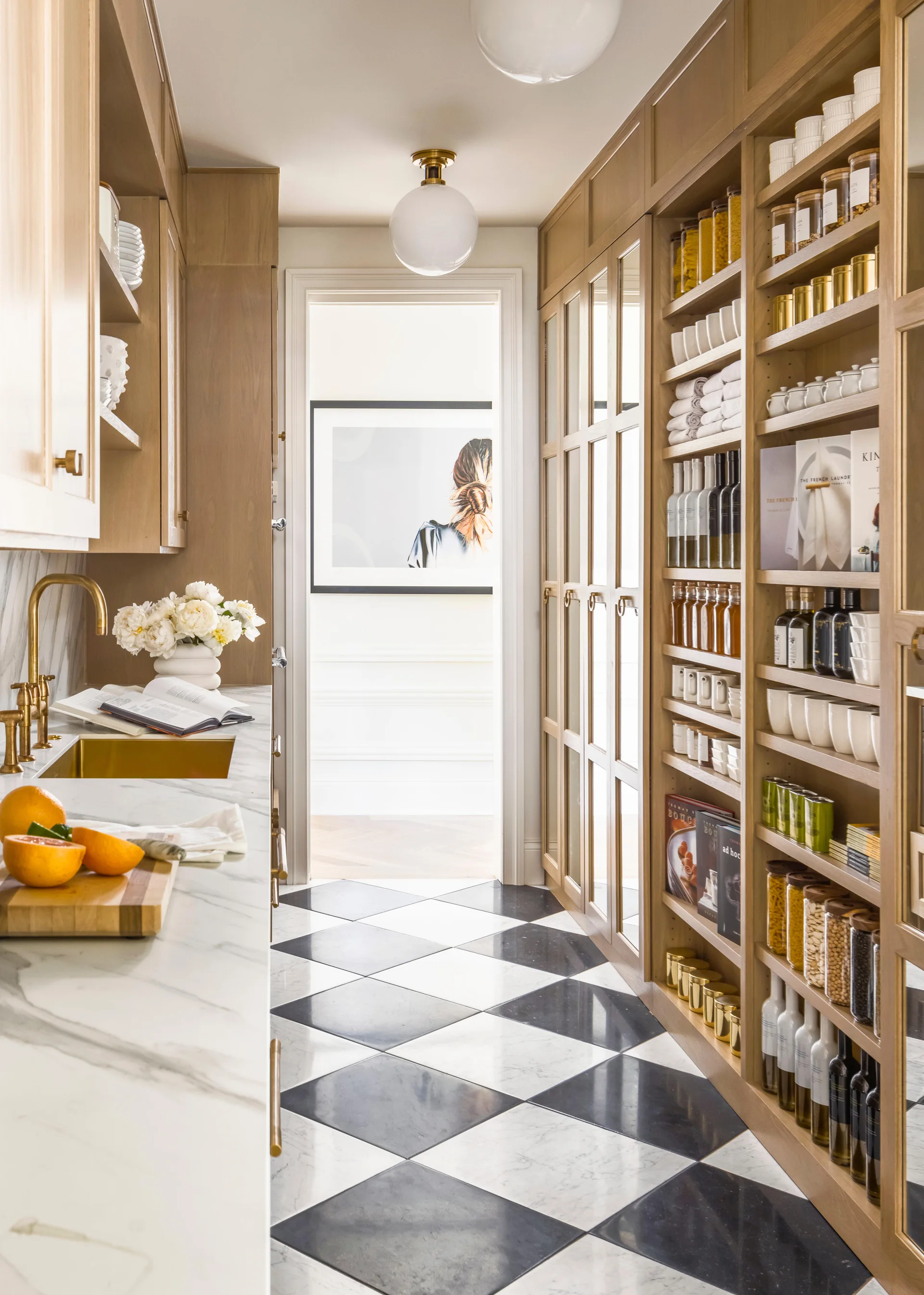 A butler's pantry features light wood built-in shelving, a marble countertop with a gold sink, and a black and white checkerboard tiled floor.